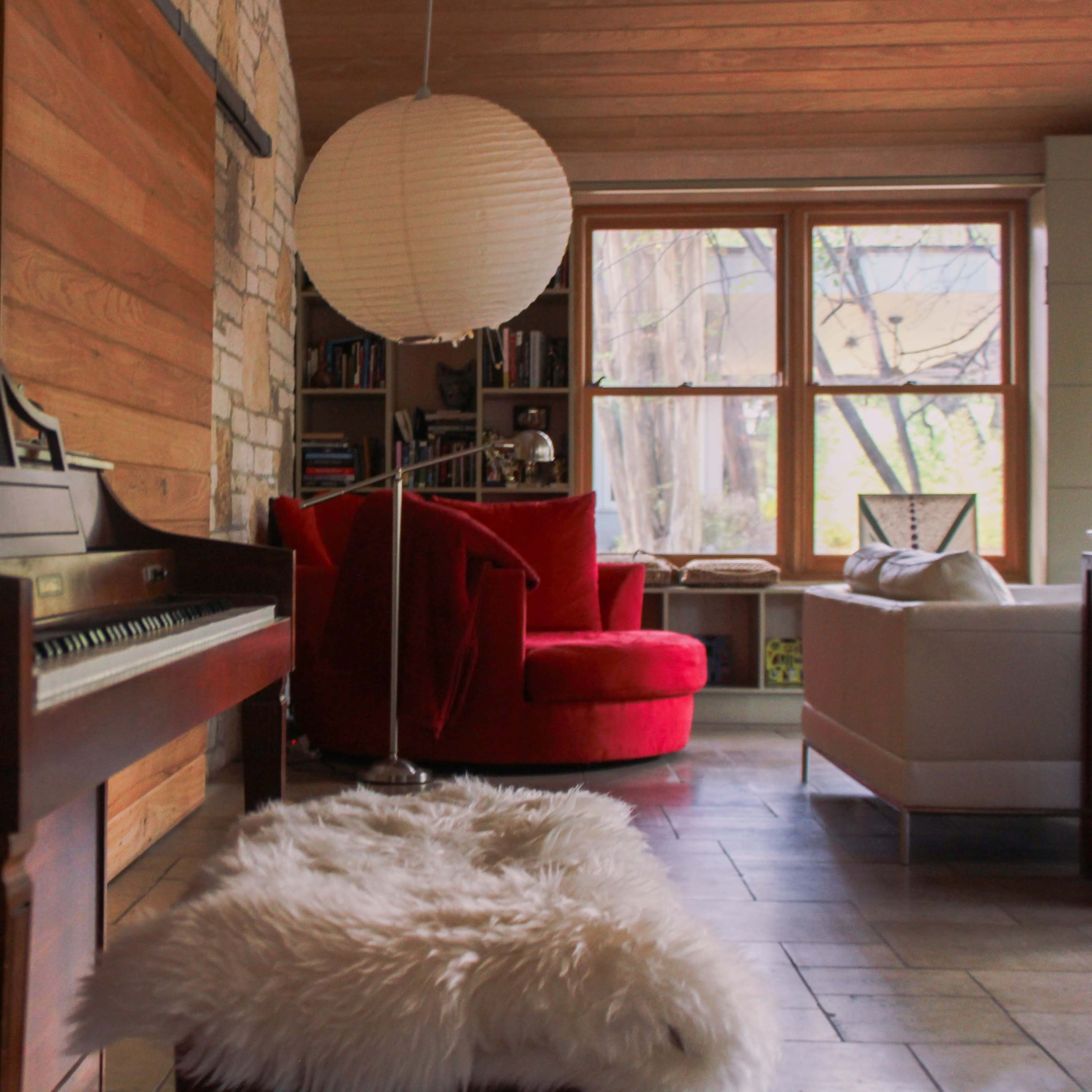 A living room featuring a wooden piano with a fur bench, a red armchair, a white sofa, and a large round paper lantern hanging above.
