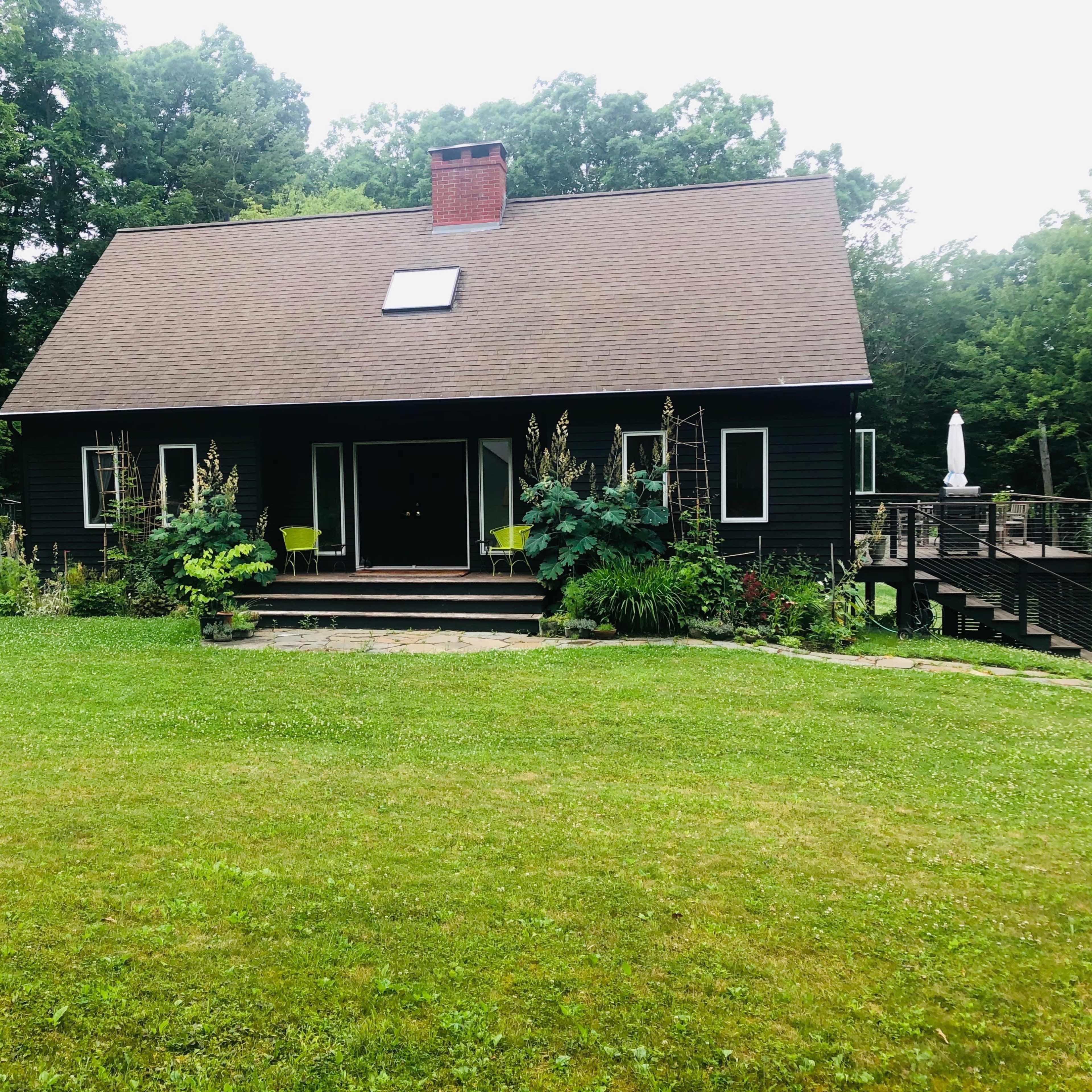 A black, two-story house with a steep roof and a front yard surrounded by trees is situated on a grassy area.