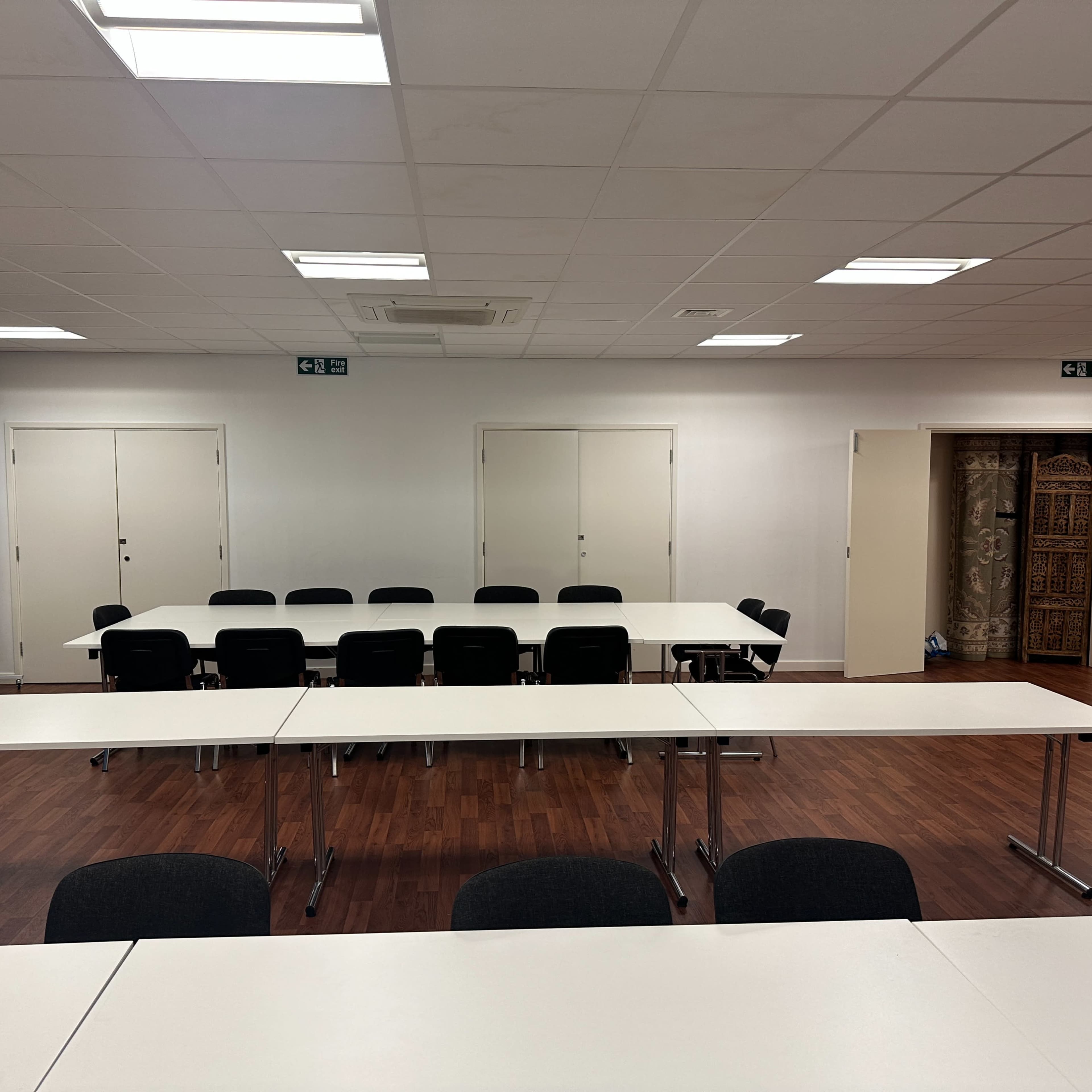 A meeting room features multiple tables arranged in a U-shape with chairs, whiteboards on the walls, and closed doors at one end.