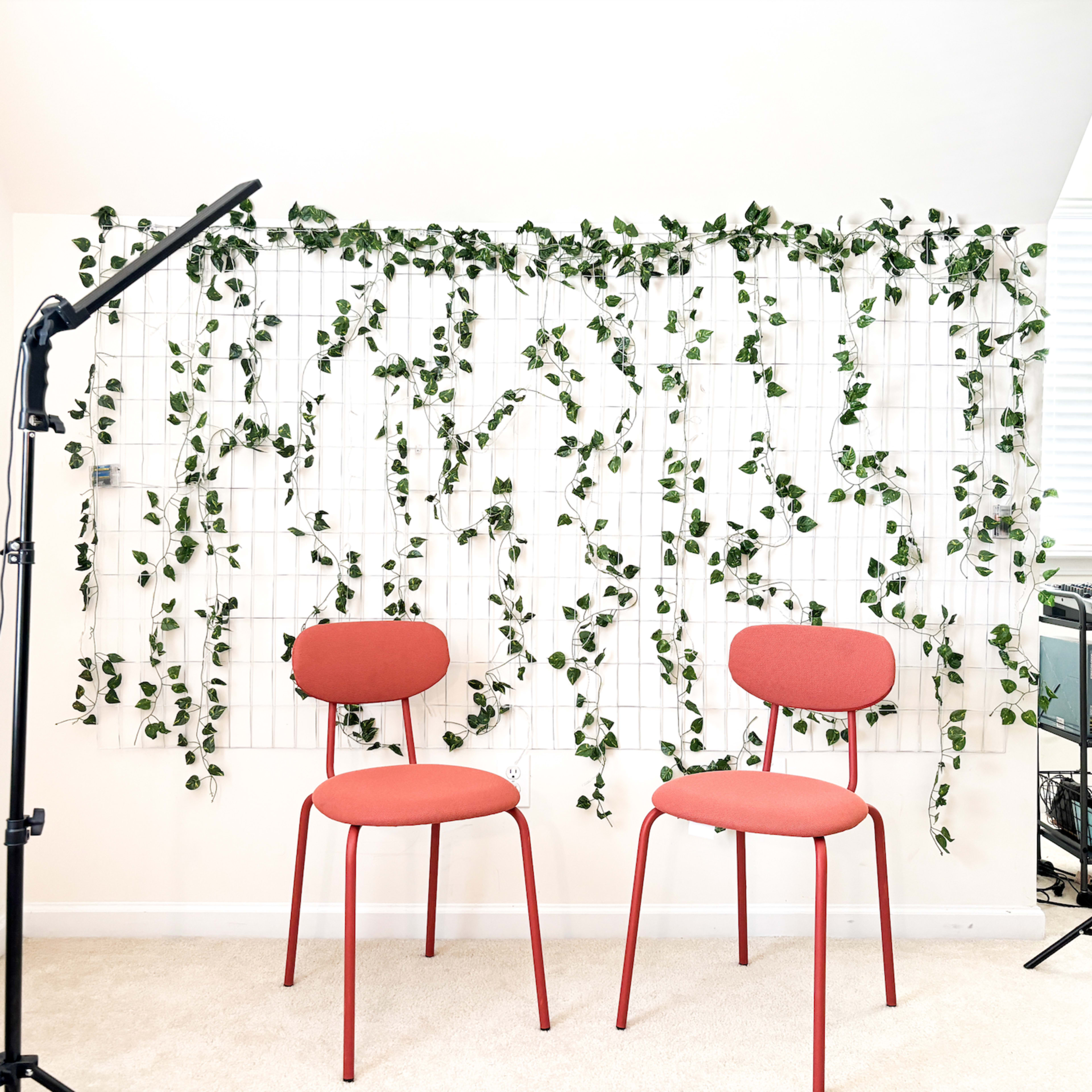 Two red chairs are positioned in front of a wall covered with green vines, illuminated by a floor lamp.