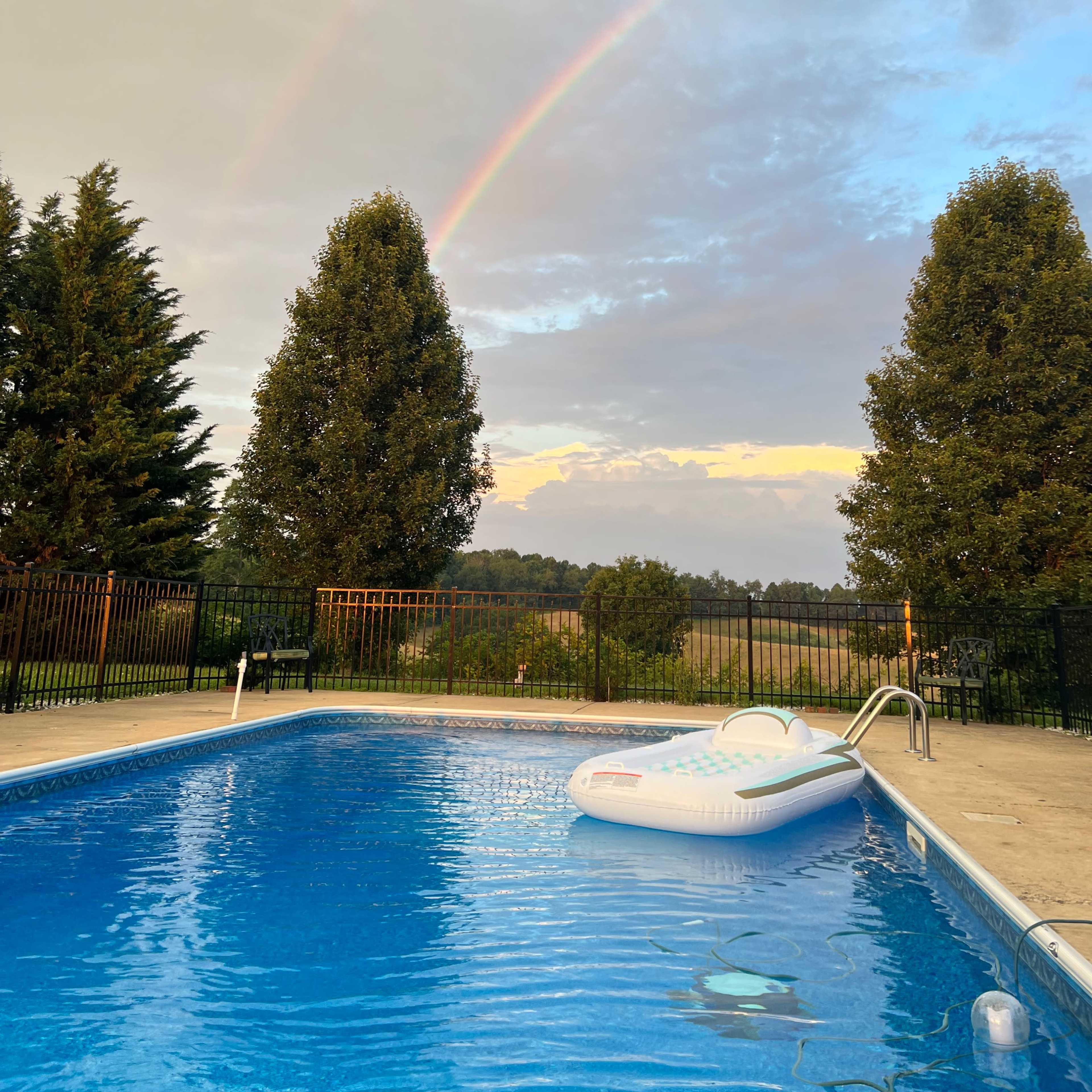 A swimming pool with an inflatable float sits beneath a double rainbow and a cloudy sky.