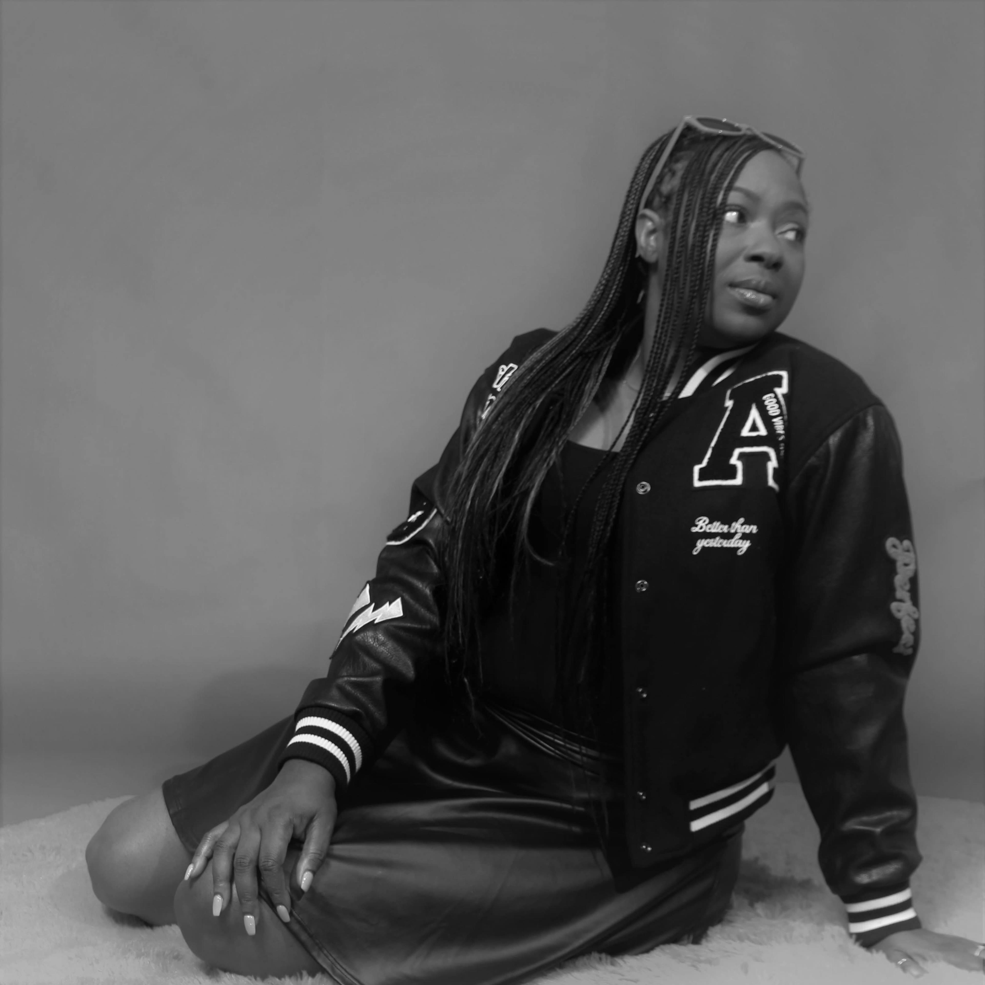 A person in a black leather jacket and skirt is sitting on a soft surface while looking over their shoulder.