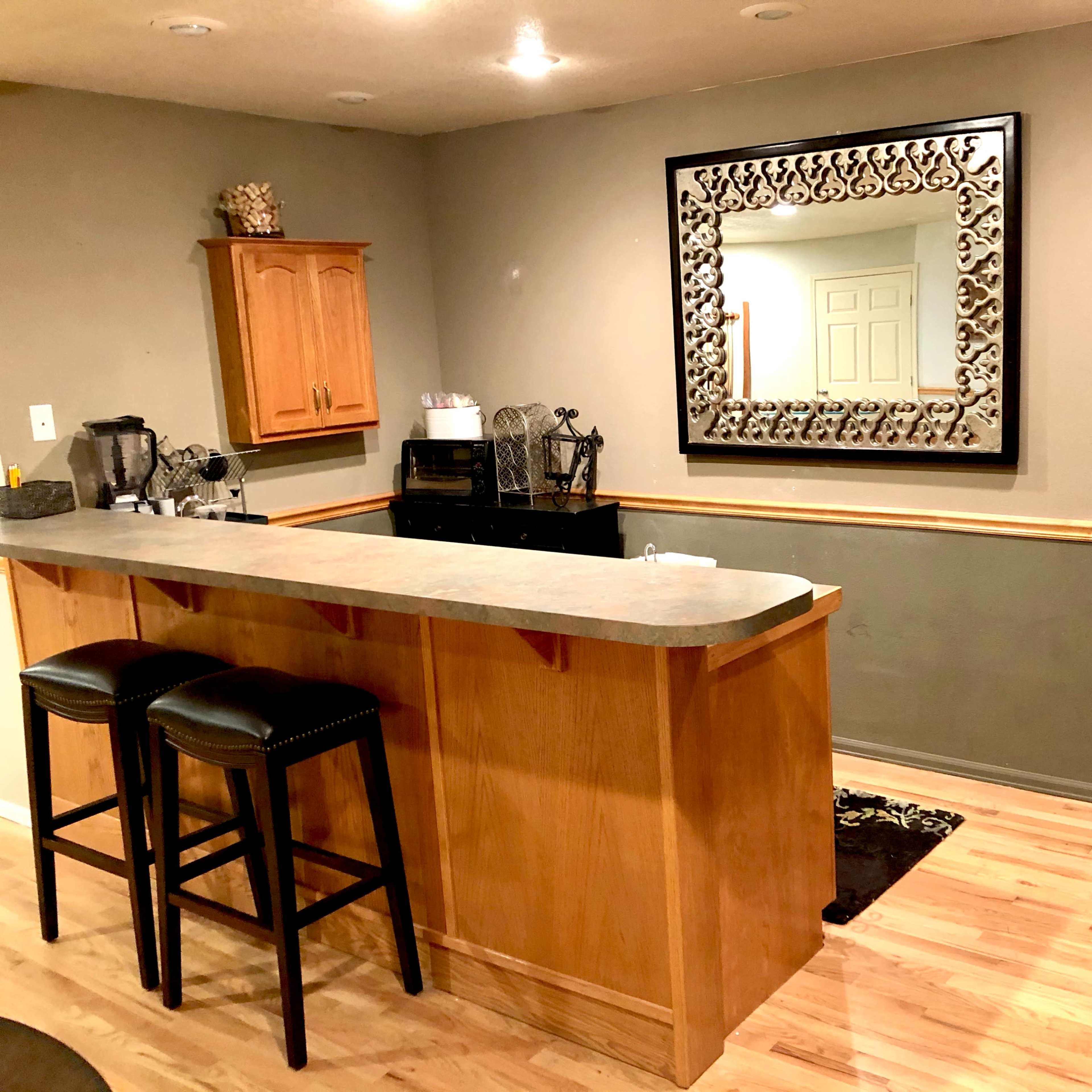 A simple kitchen area with a bar counter, stools, and a stainless steel refrigerator, featuring a mirror on the wall and wooden cabinetry.