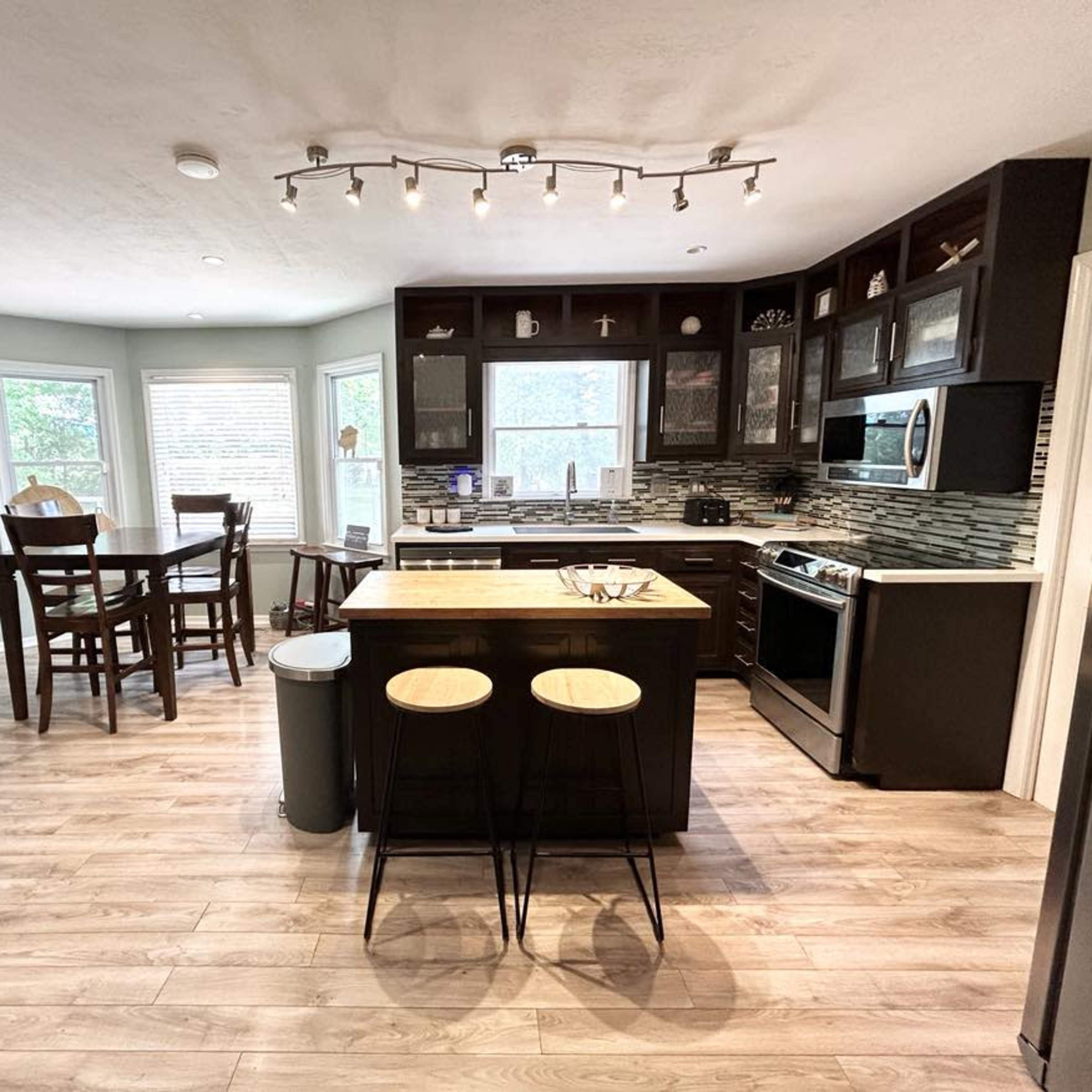 A modern kitchen featuring an island with two stools, a dining table, and large windows that provide natural light.