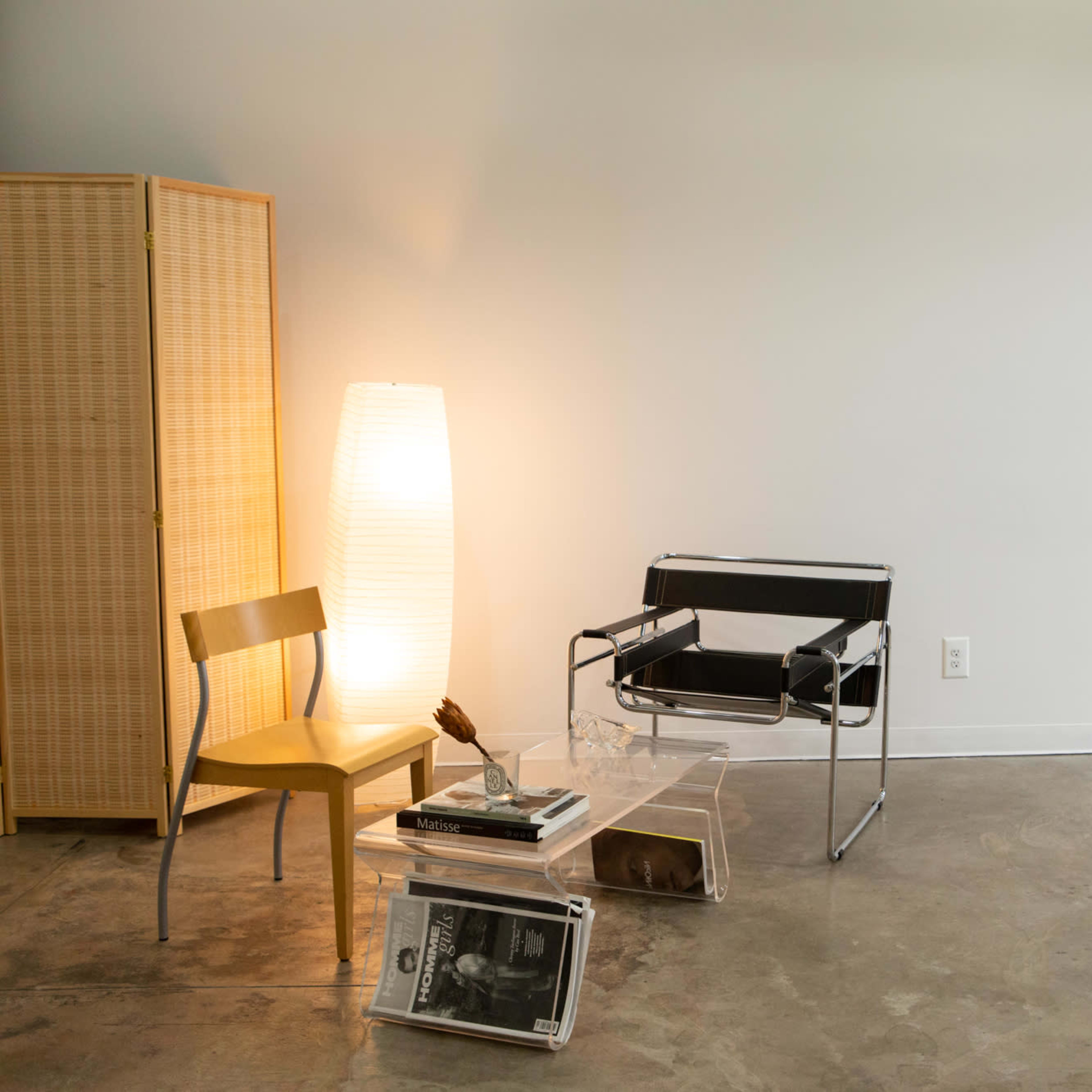 A minimalist interior with a black and chrome chair, a wooden chair, a tall lamp, and a small side table stacked with magazines, set against a light-colored wall.