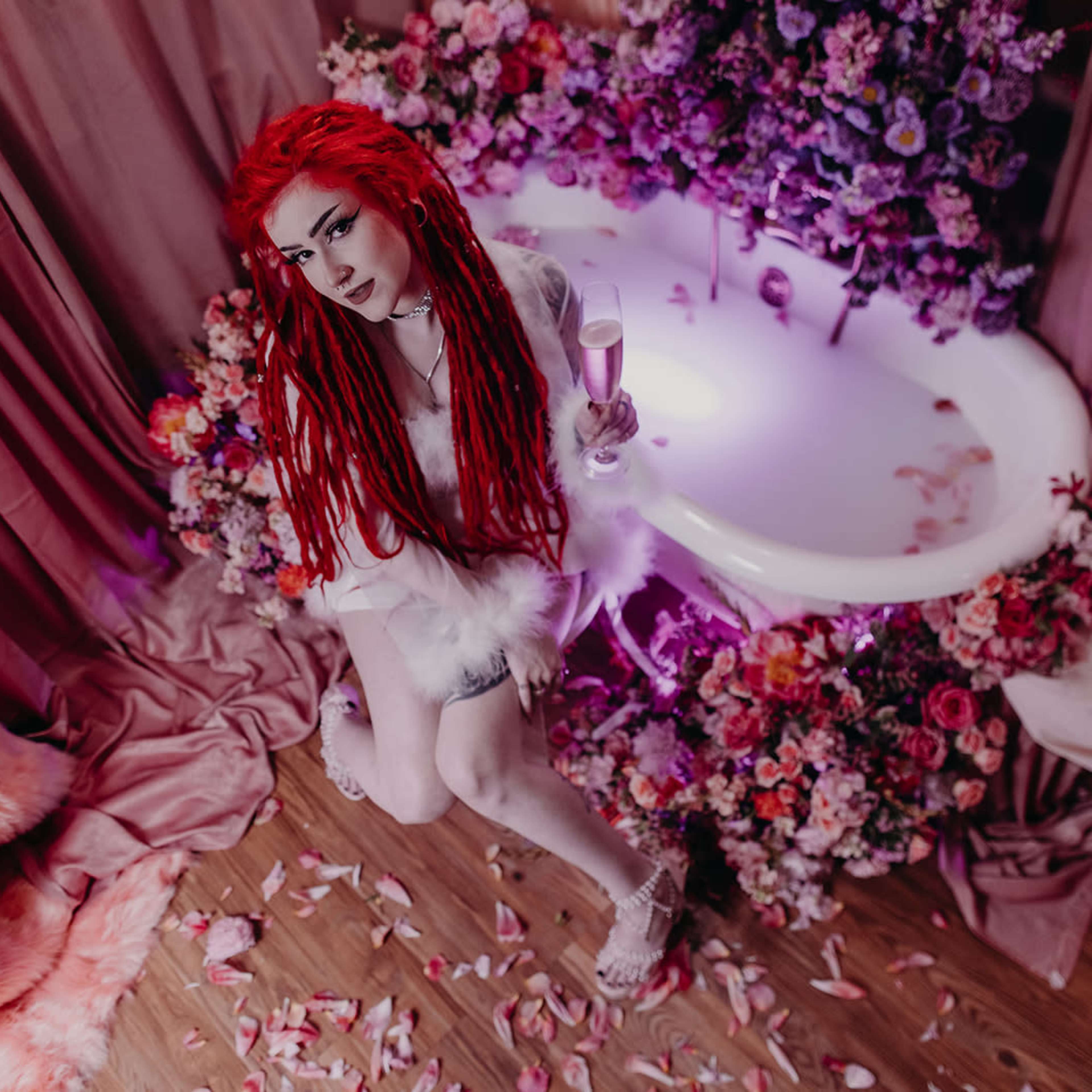 A person with long red hair sits beside a flower-adorned bathtub surrounded by petals and draped fabrics.