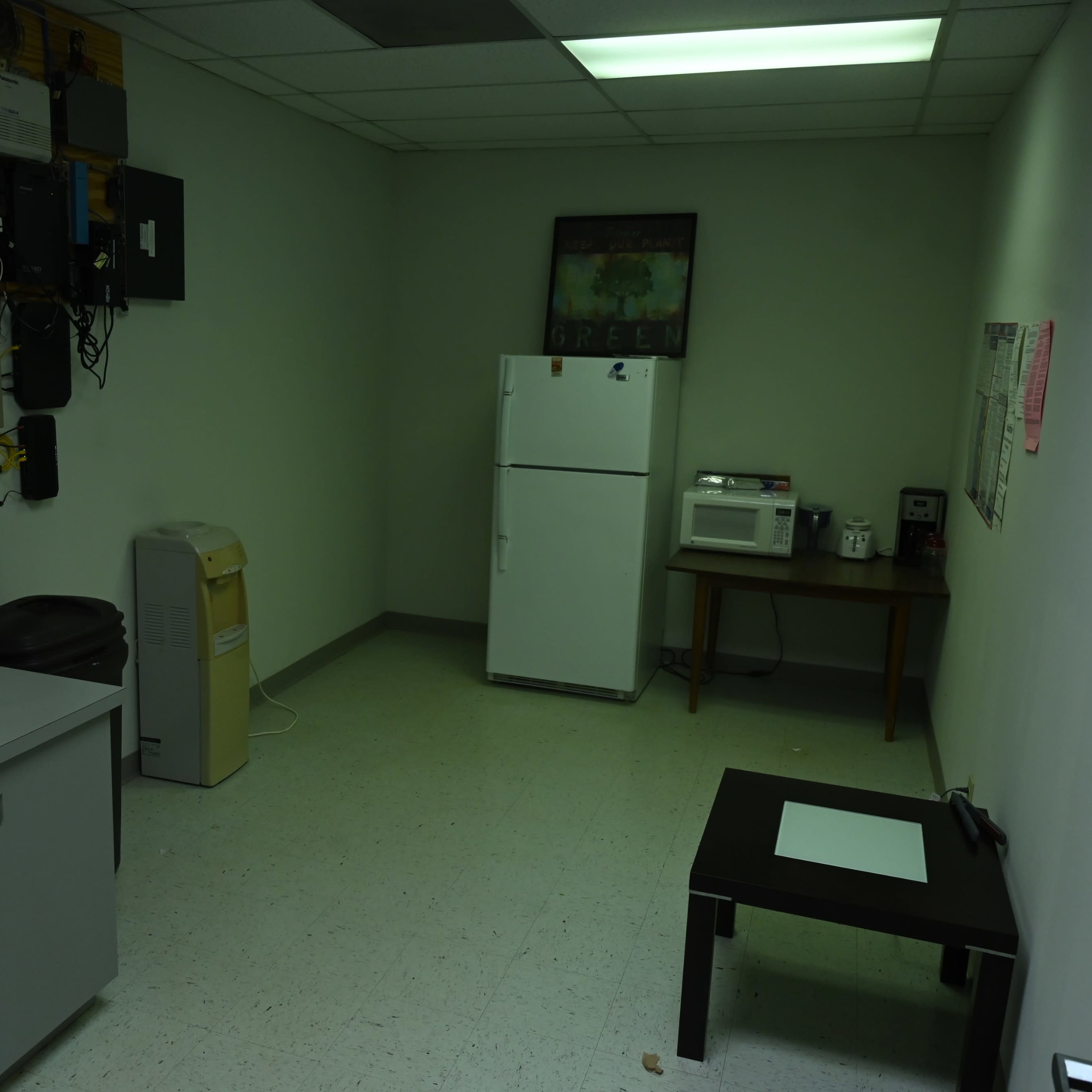 A sparsely furnished break room with a white refrigerator, a table, a microwave, and a waste bin against plain walls.