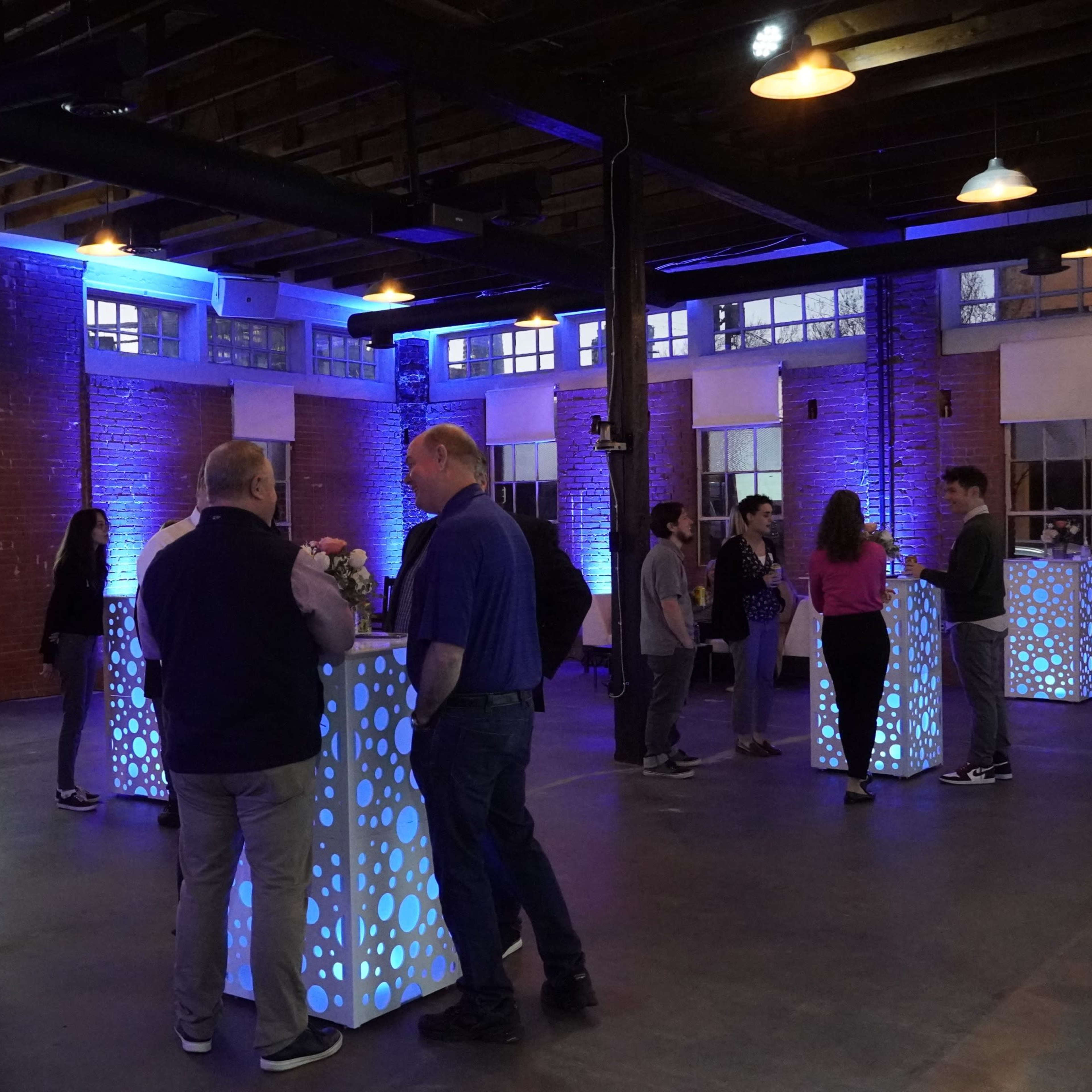 A group of people mingles in a well-lit event space with illuminated tables and exposed brick walls.