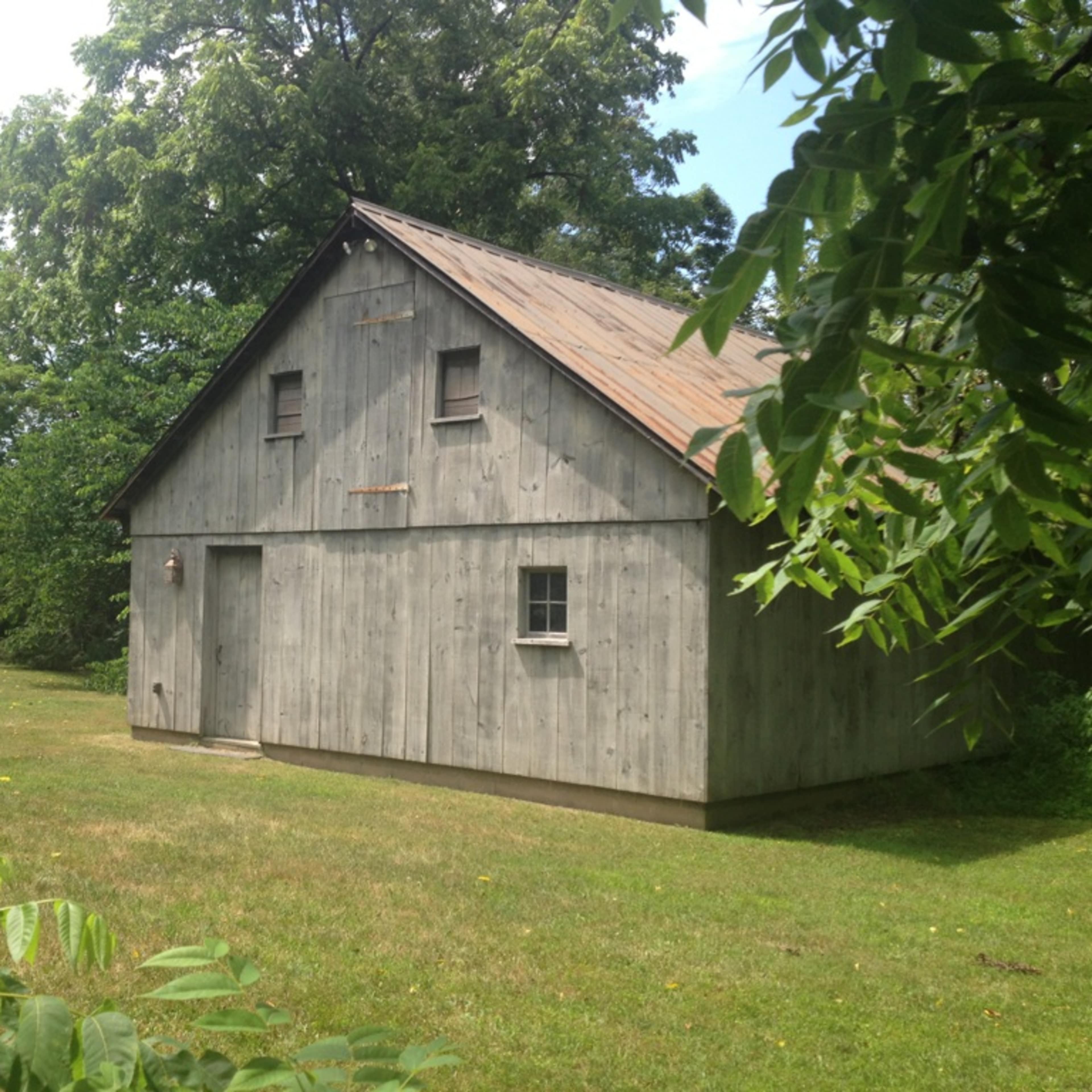 A weathered wooden shed with a corrugated metal roof, set on a grassy area surrounded by trees.