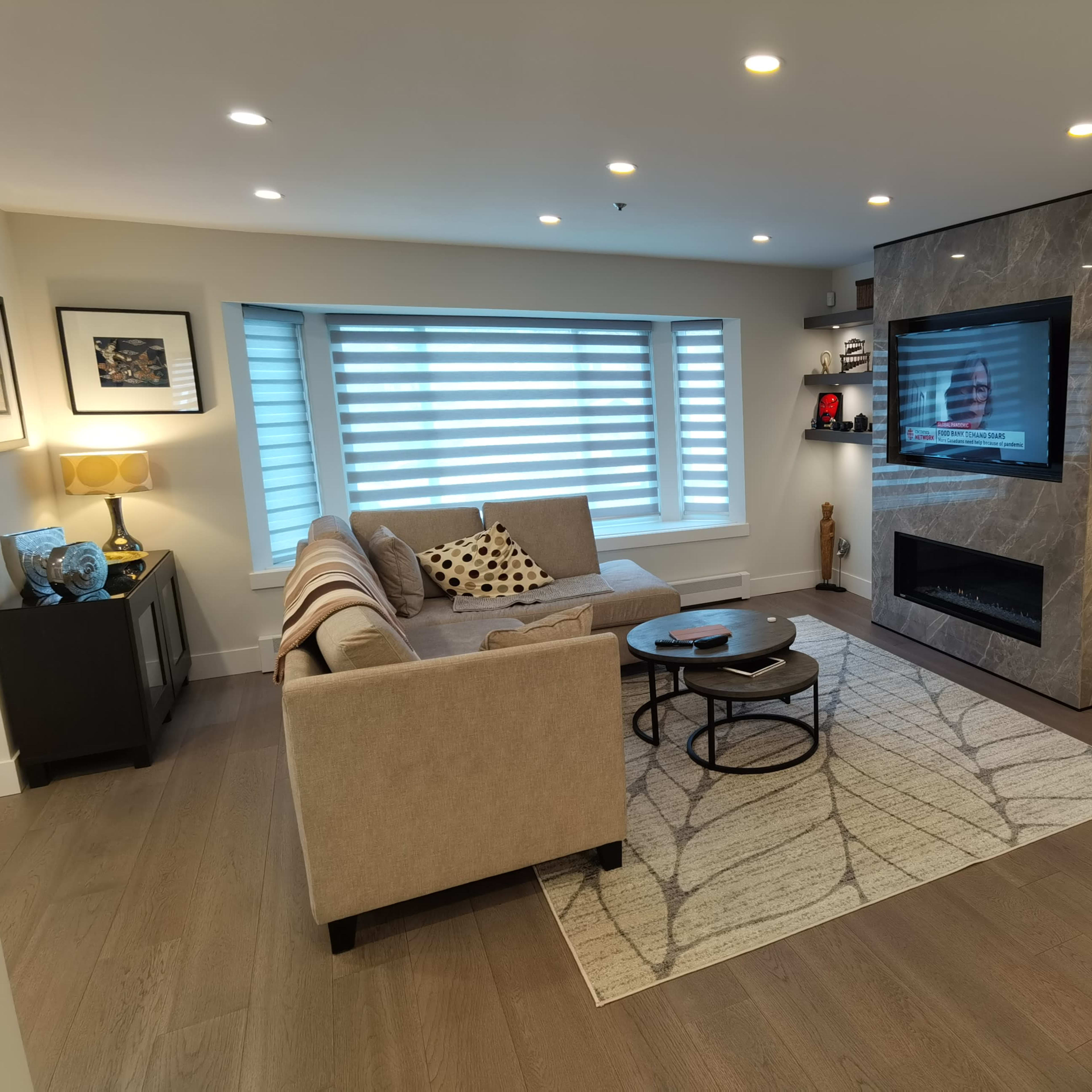 The living room features a sectional sofa, a round coffee table, a wall-mounted TV, and decorative shelves, with soft lighting and a large area rug.