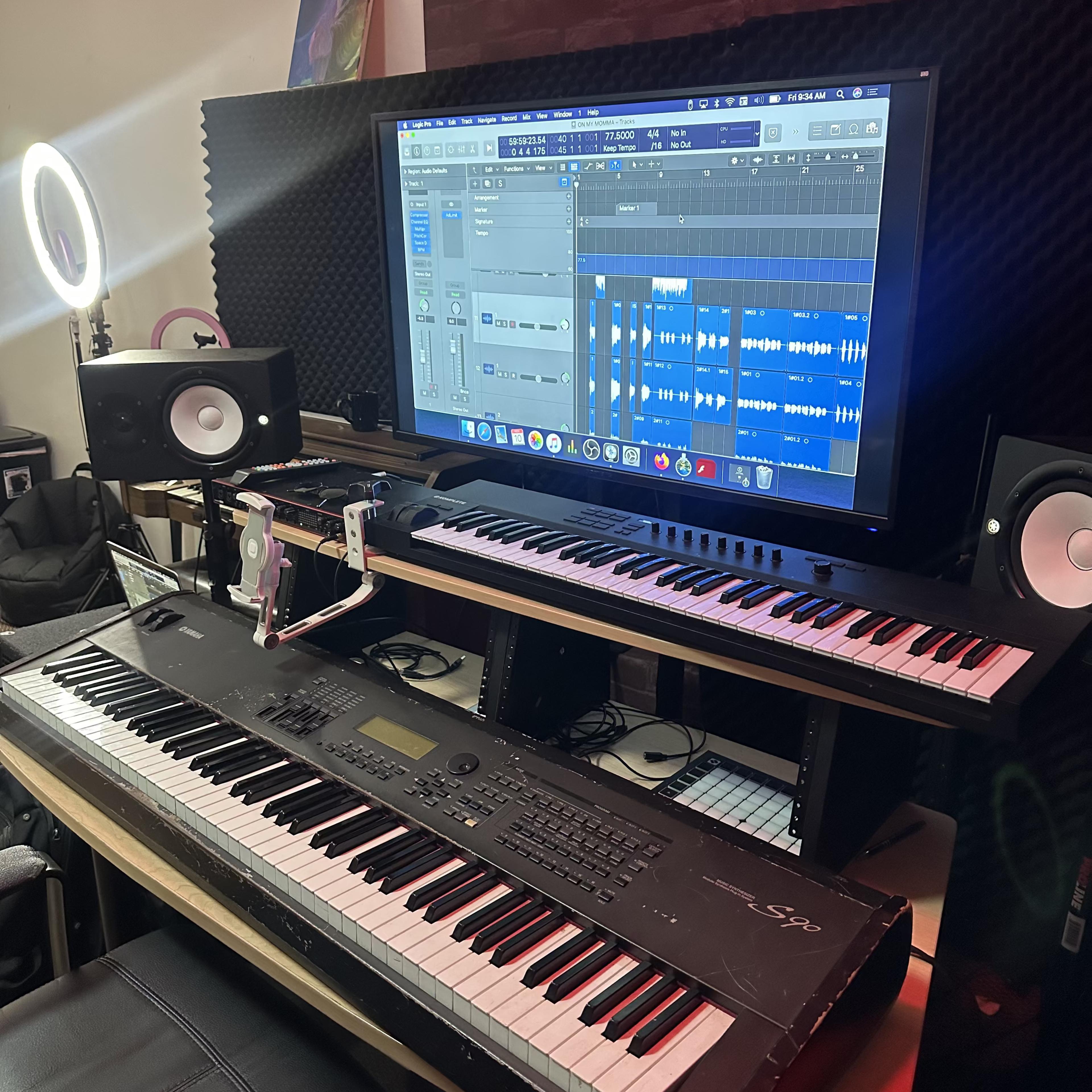 A music production setup features a digital audio workstation displayed on a large monitor, with two keyboards and studio monitors on a desk.