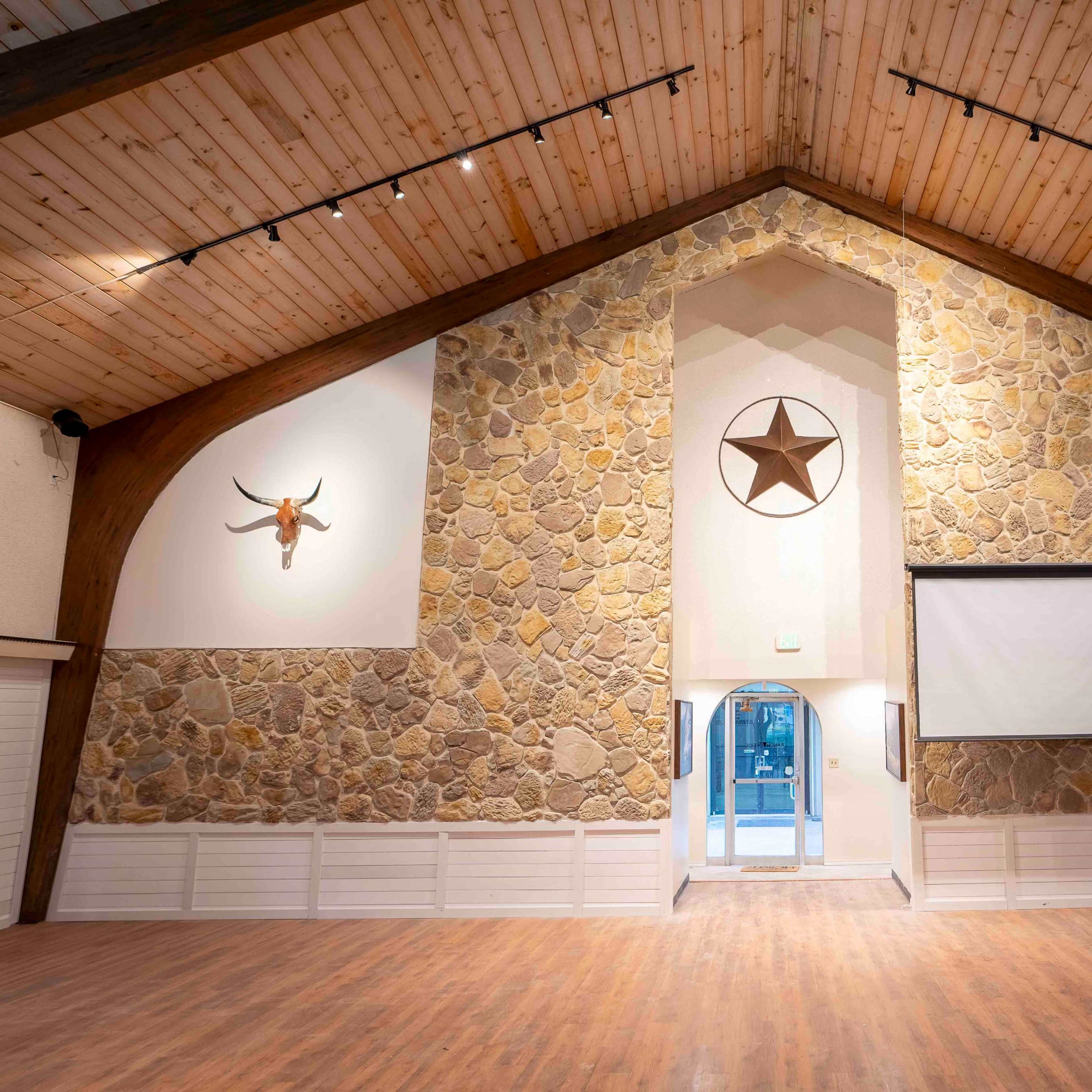 The interior of a spacious hall features a stone wall with a large star emblem, wooden beams, and two mounted horns on either side.