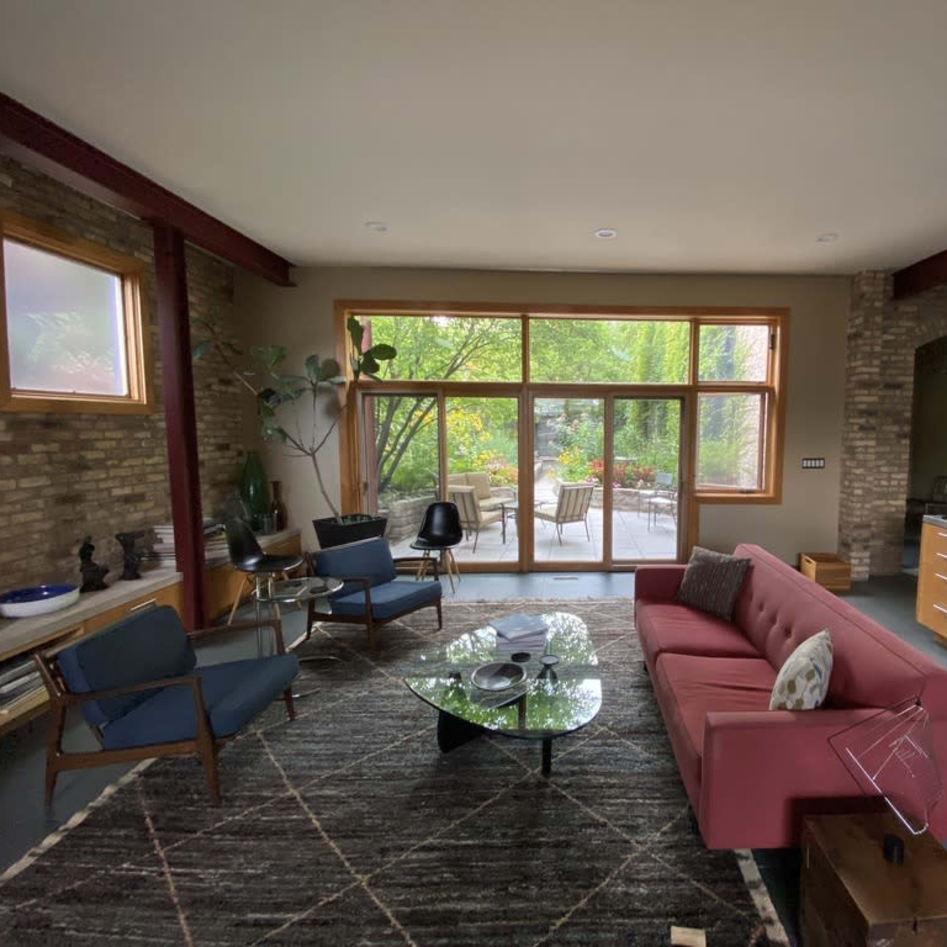 A modern living room with a red sofa, a glass coffee table, and large windows overlooking a green outdoor area.