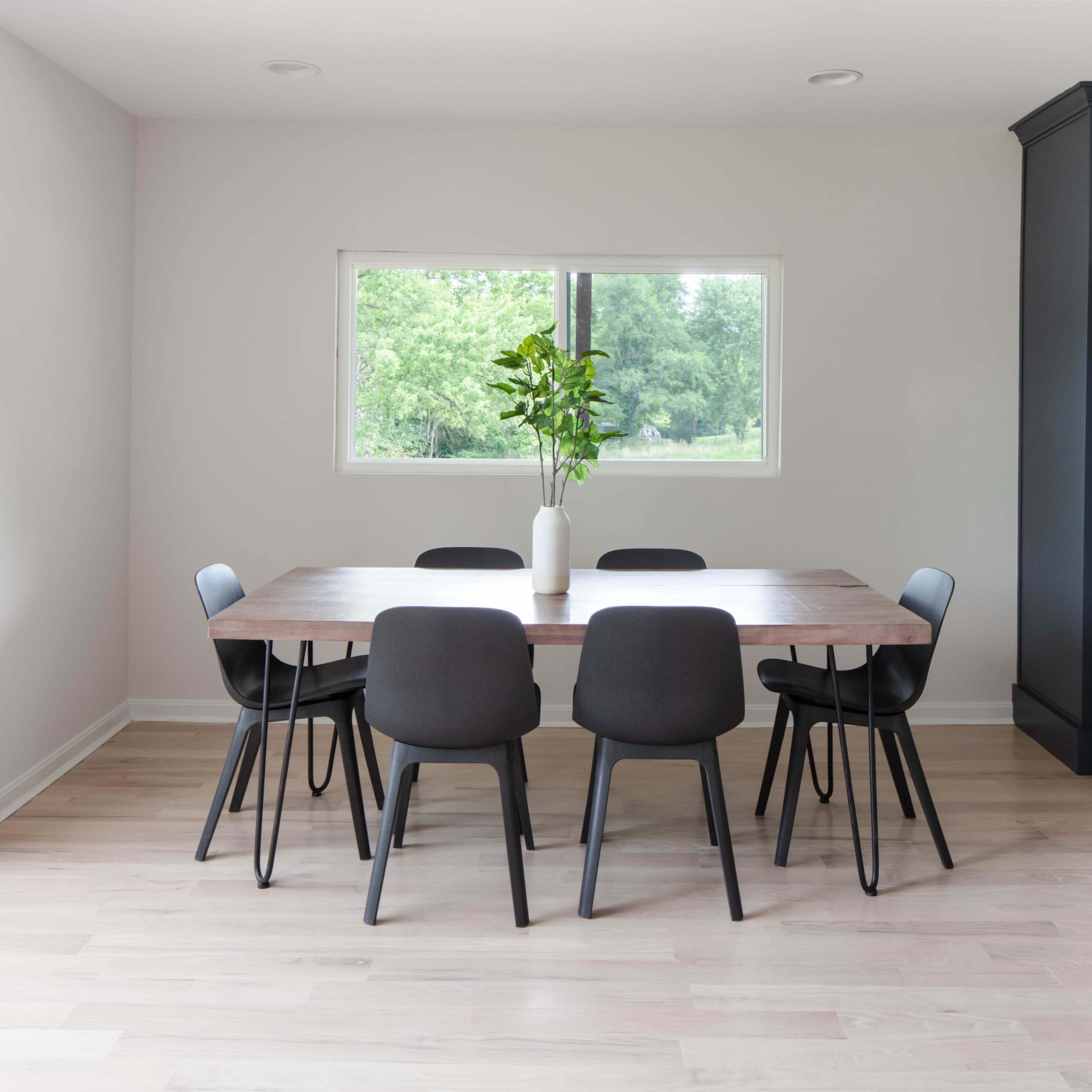 A modern dining area features a wooden table surrounded by six black chairs, with a vase of greenery as the centerpiece and a large window offering a view of the outdoors.