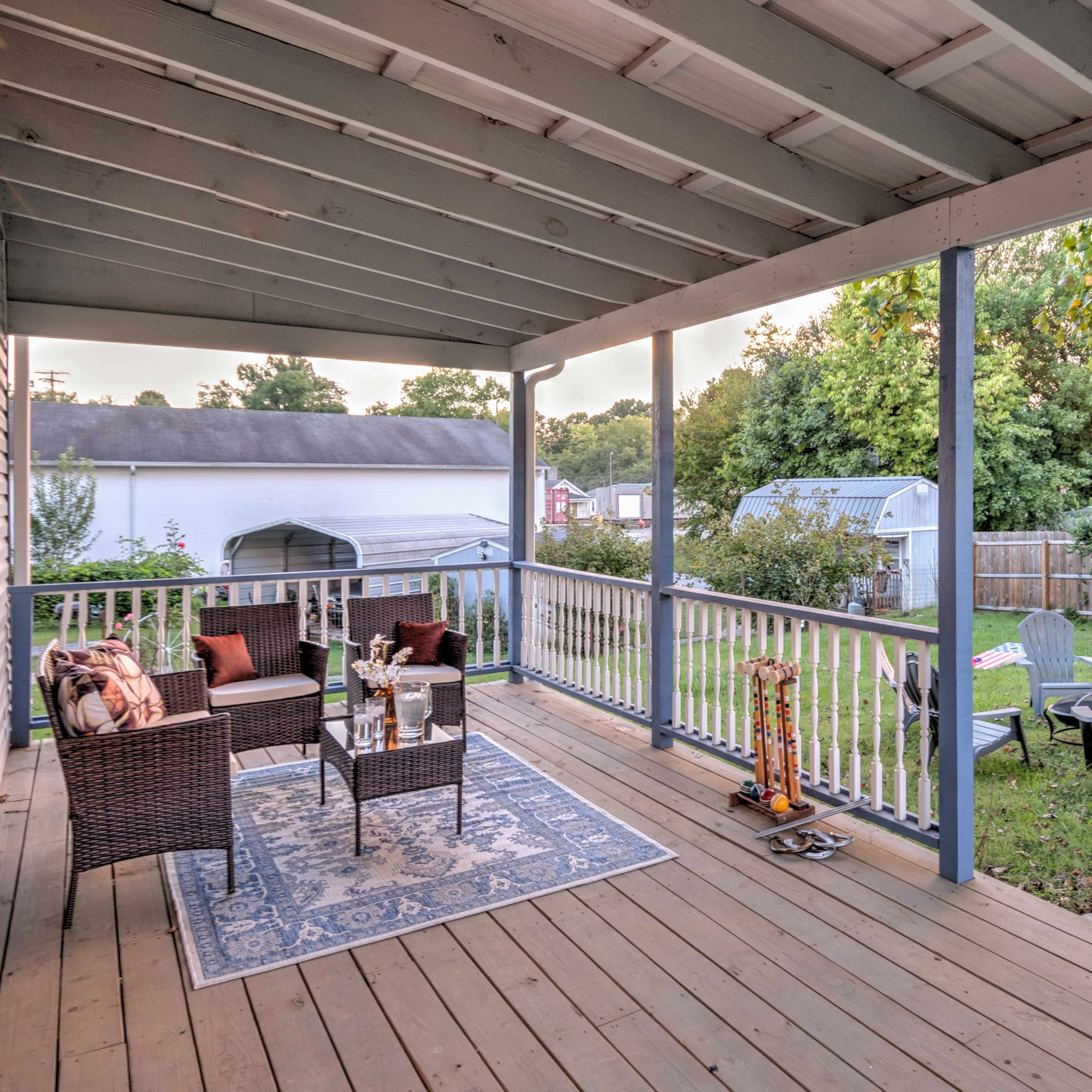 A covered porch with two wicker chairs and a small table overlooks a grassy yard with several chairs and a shed in the background.