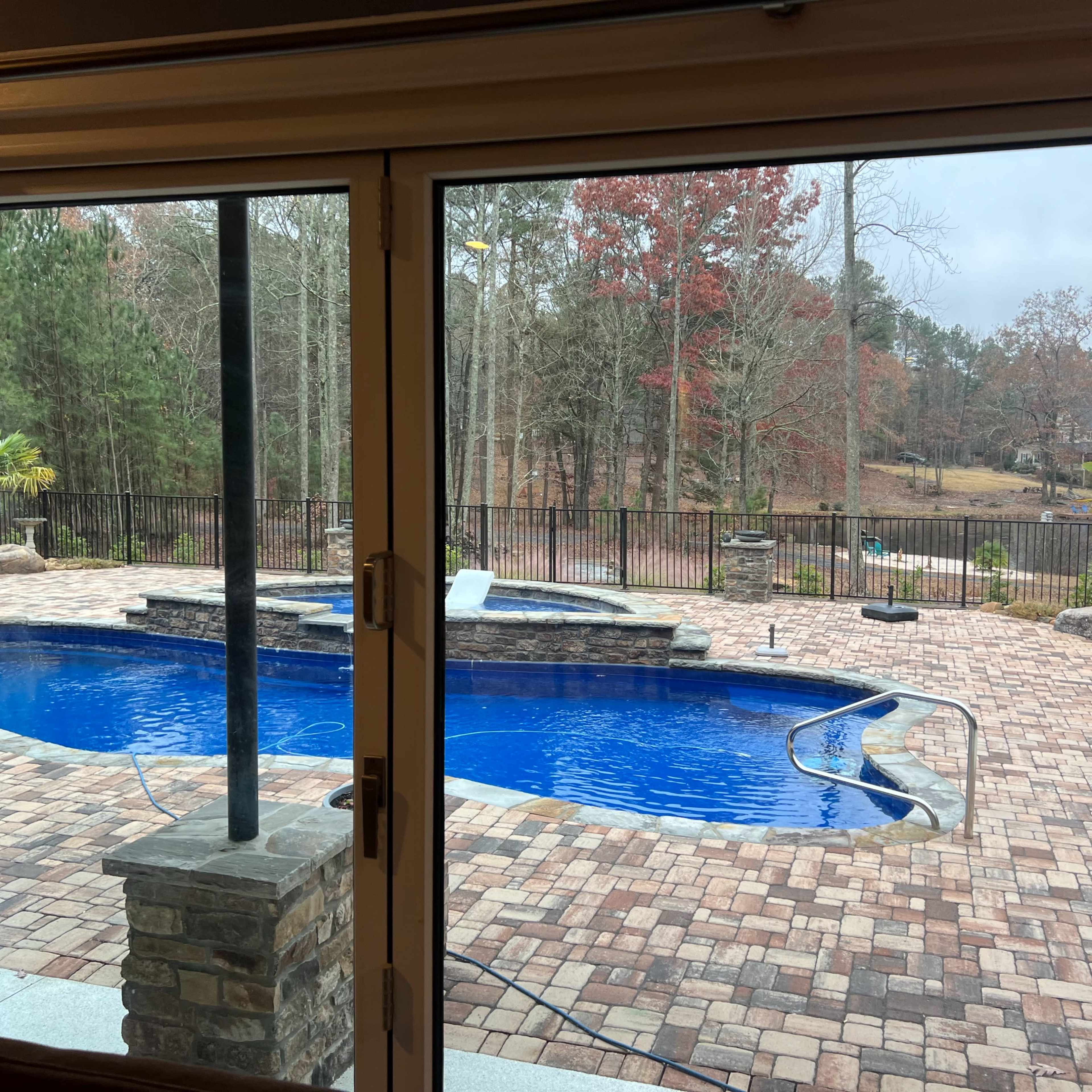 A view from inside a house shows a landscaped backyard with a blue swimming pool surrounded by a brick patio and a fenced area.