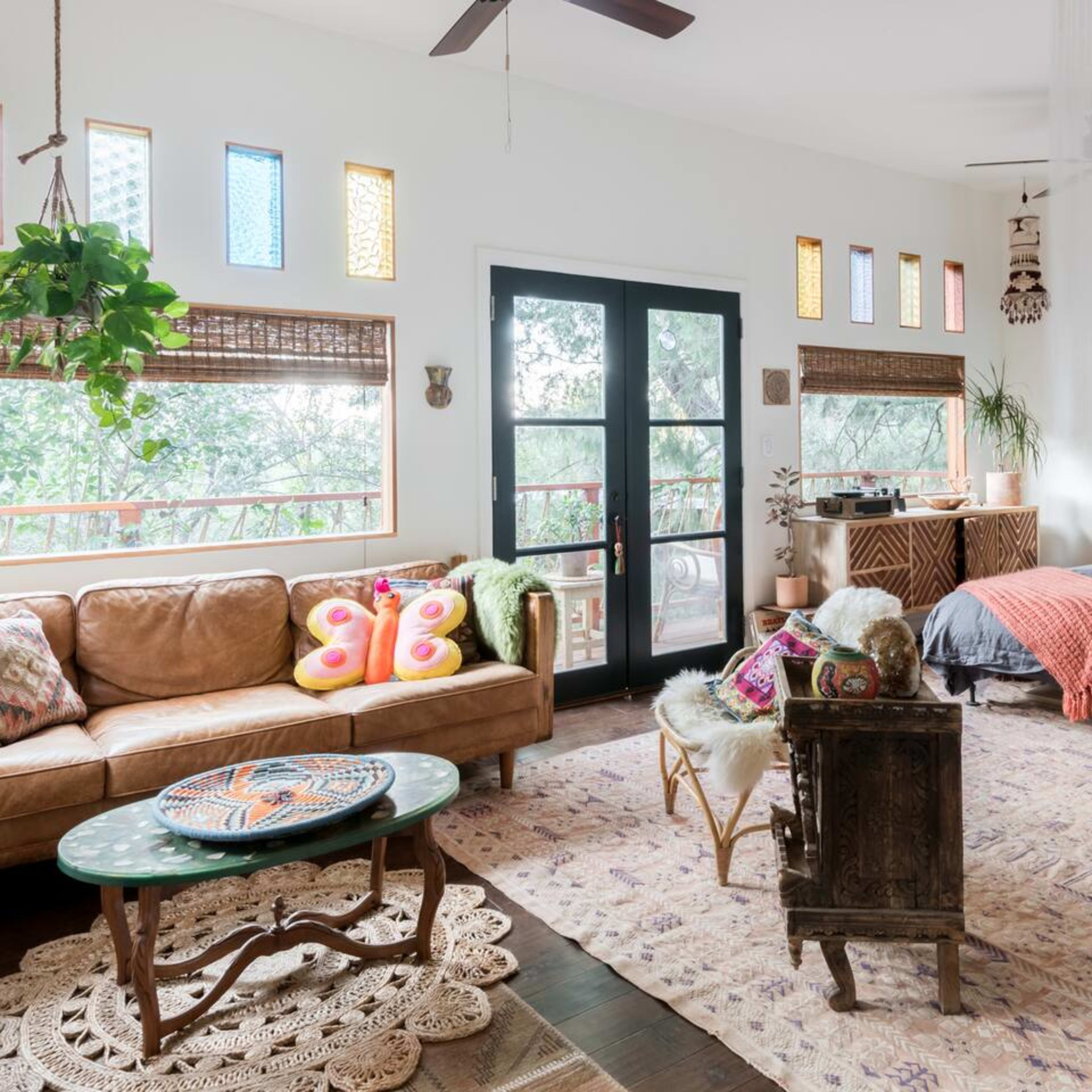 A cozy living room furnished with a leather sofa, colorful pillows, a circular table, and decorated with plants and natural light filtering through patterned windows.