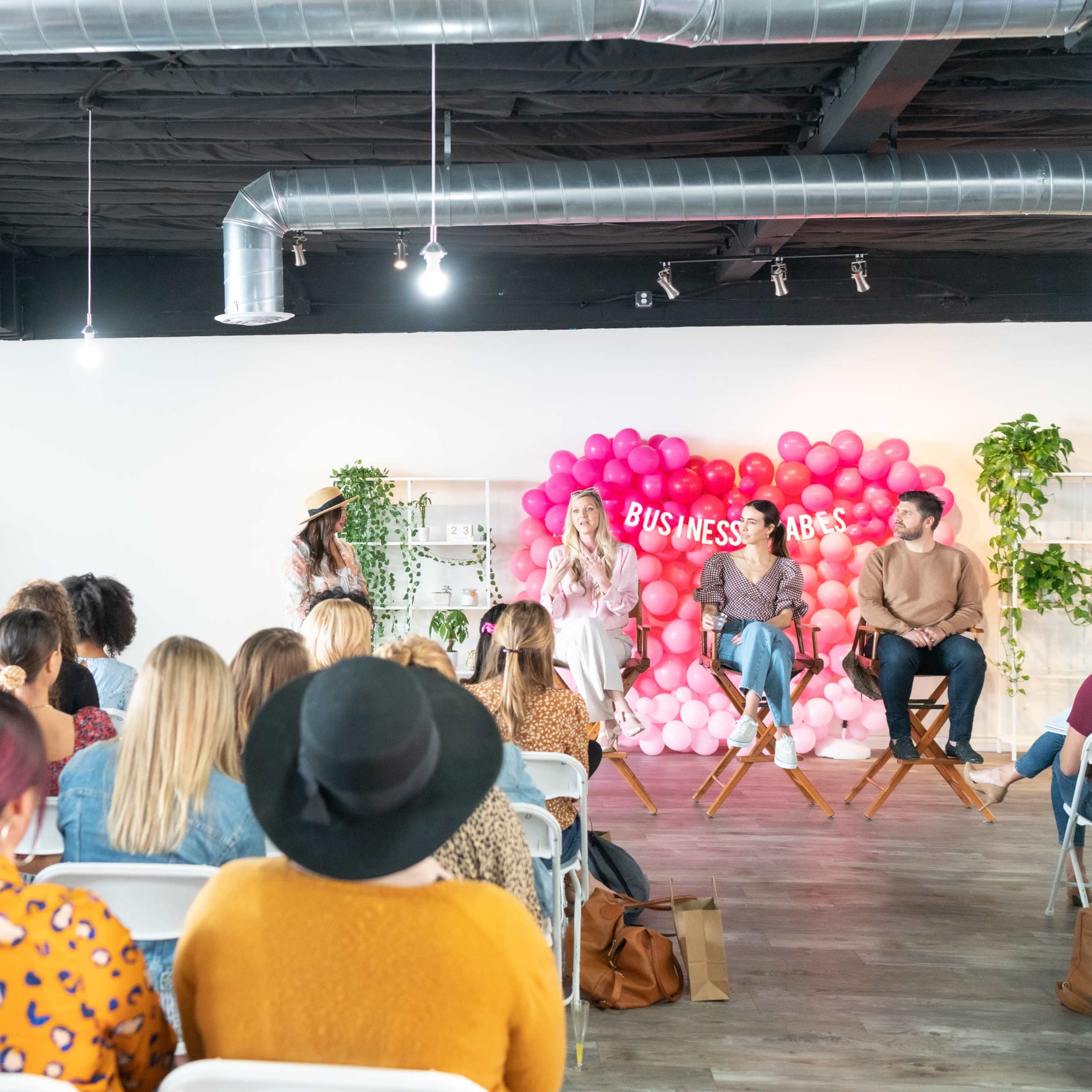 A group of people sit in a conference room, facing a panel discussion staged against a backdrop of pink balloons.