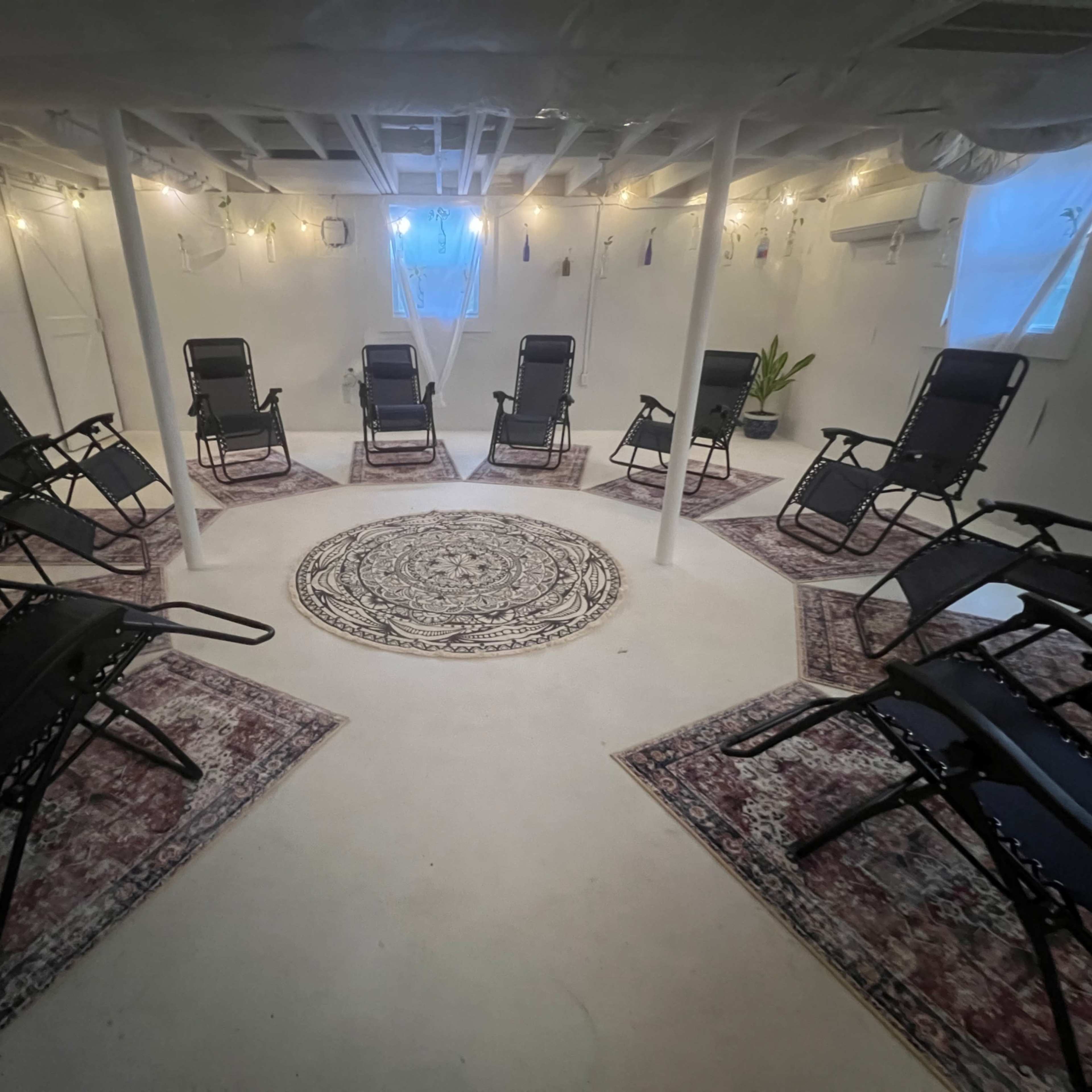 A circular seating arrangement with black reclining chairs is set on a light-colored floor adorned with a central rug in a well-lit basement space.