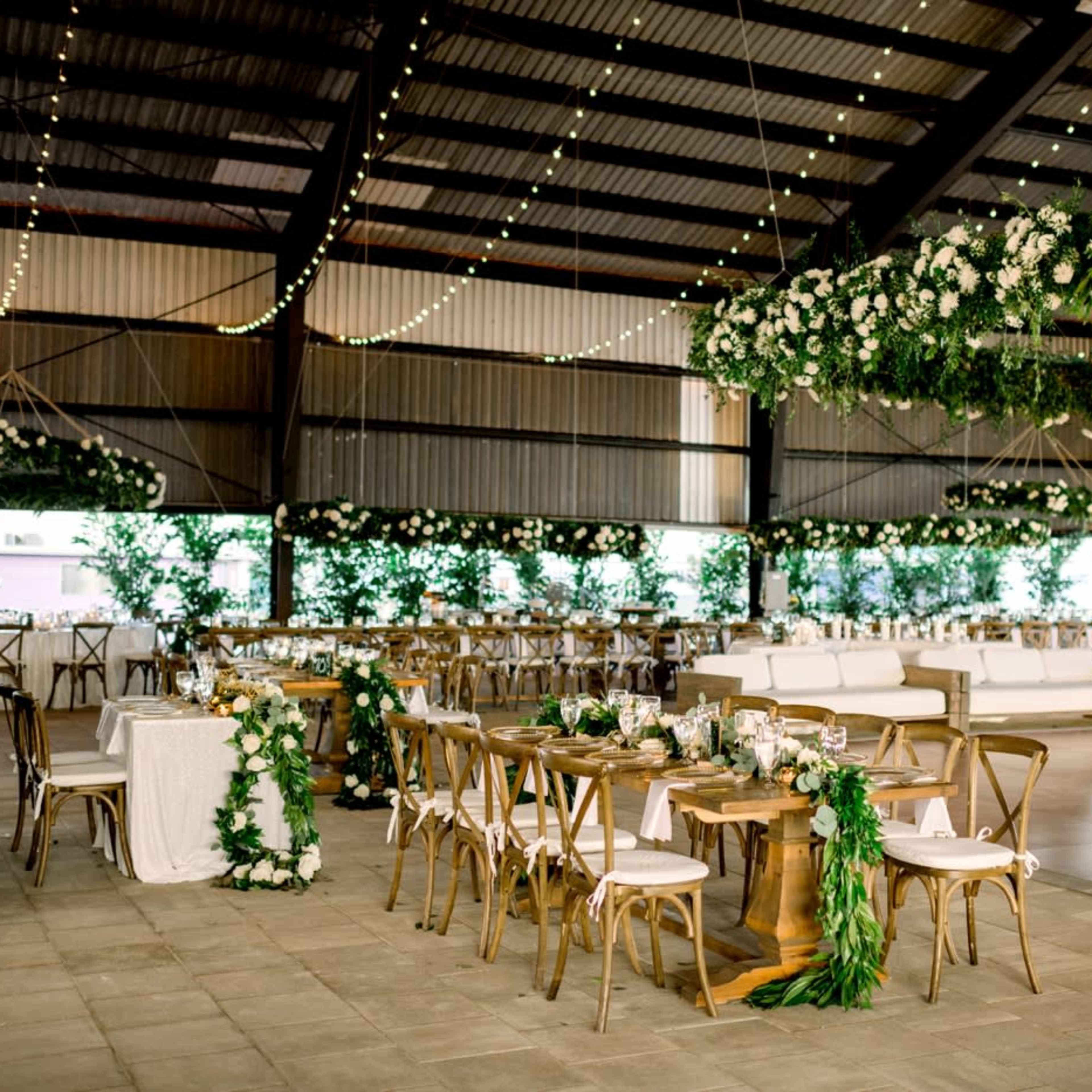 A beautifully decorated event space with wooden tables, greenery, and floral arrangements, set under a large metal structure.