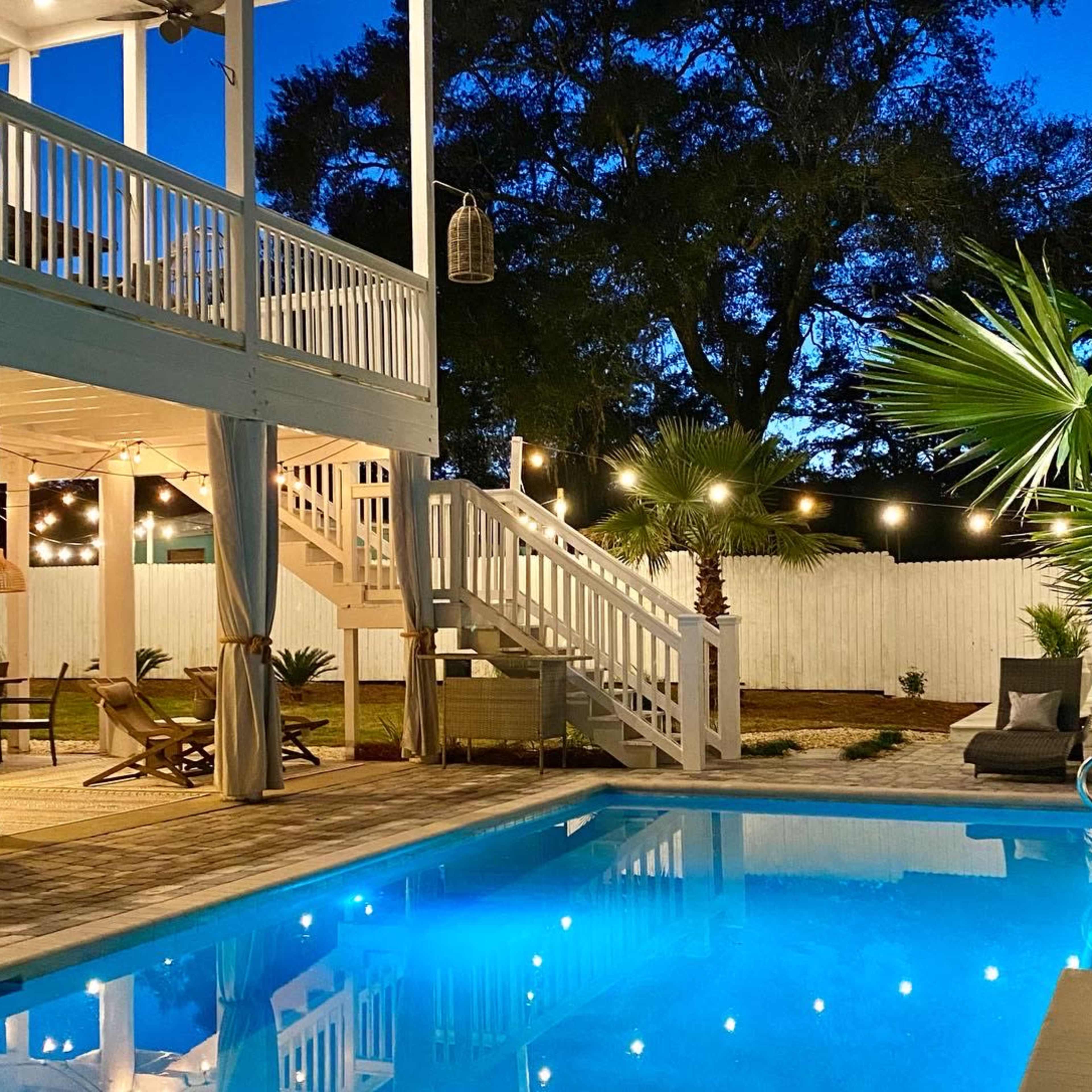 A well-lit outdoor pool area with a staircase leading to a raised deck, surrounded by palm trees and cozy seating.