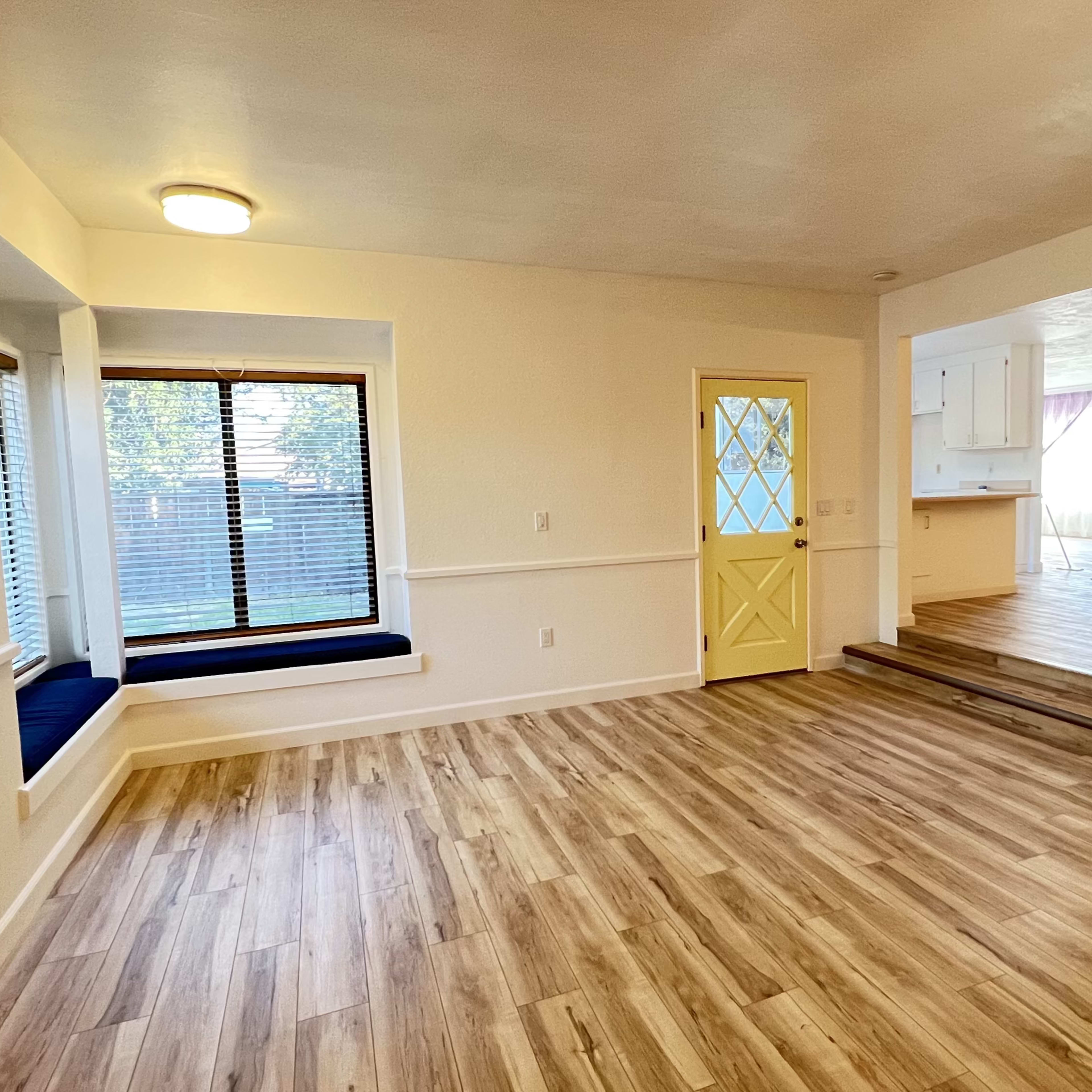 A light-filled interior space featuring a window seat, yellow door, and an open layout connecting to a kitchen area.