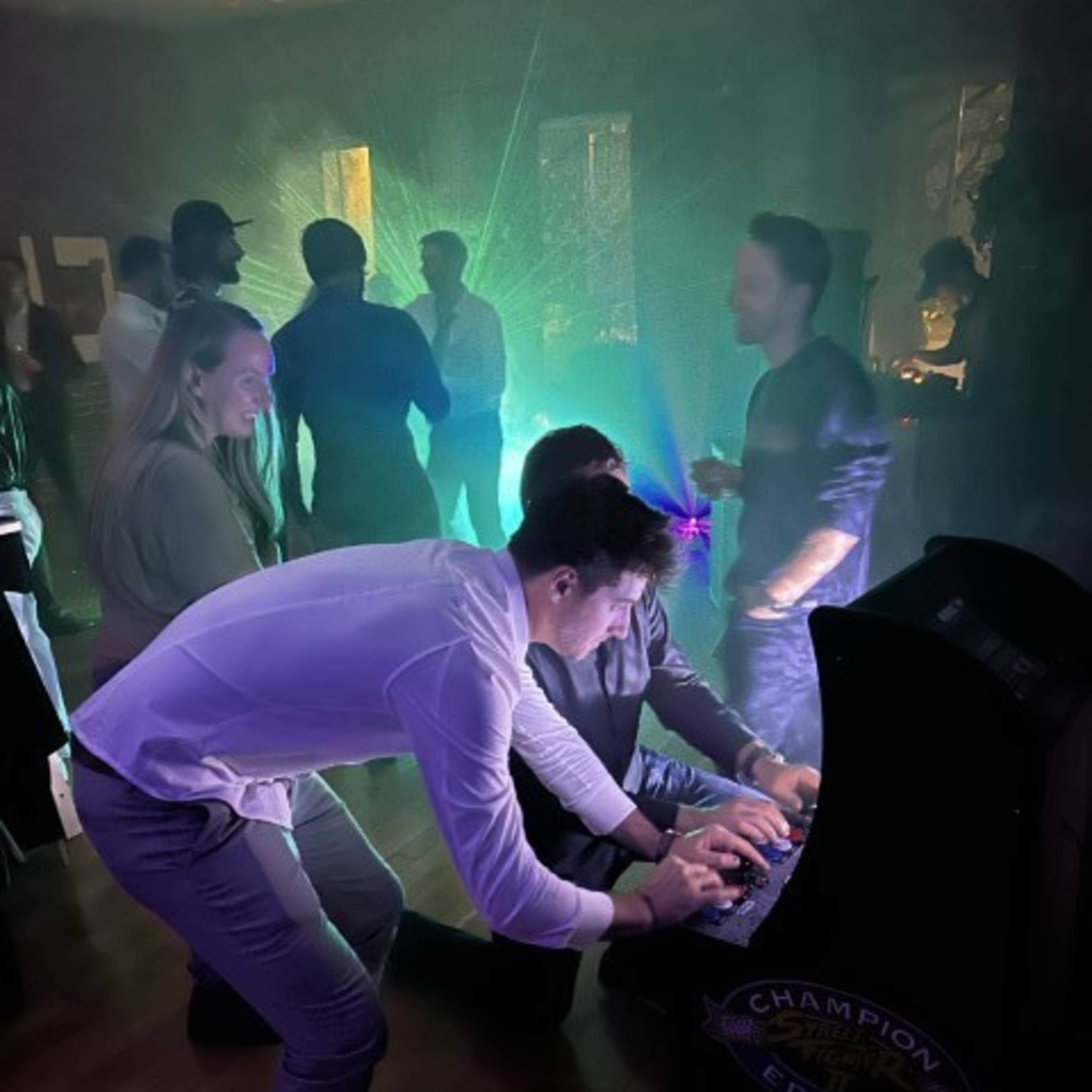 A group of people interacts around an arcade game in a dimly lit room with colorful lights.