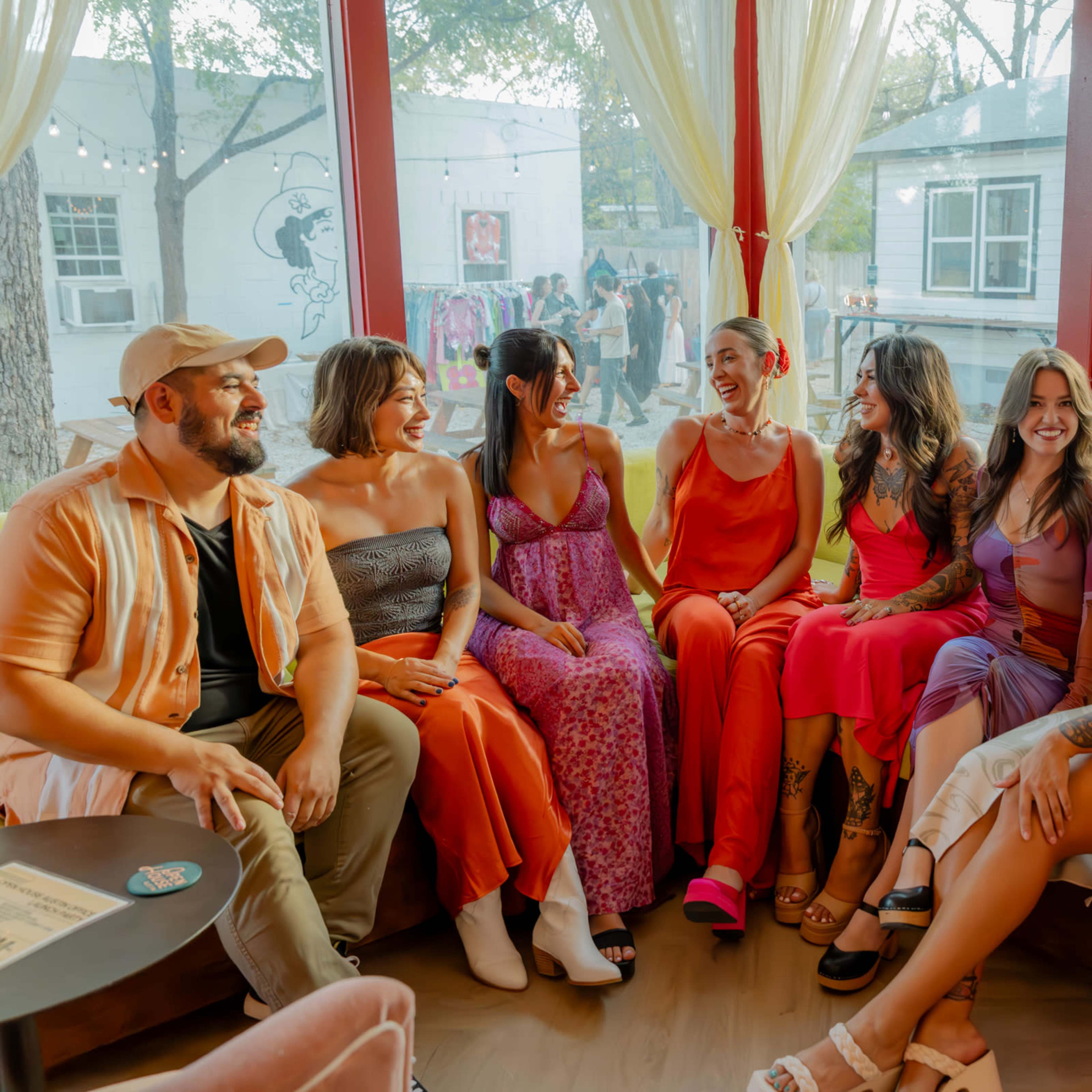 A group of eight women and one man sit together on a couch in a brightly lit room, smiling and interacting with each other.