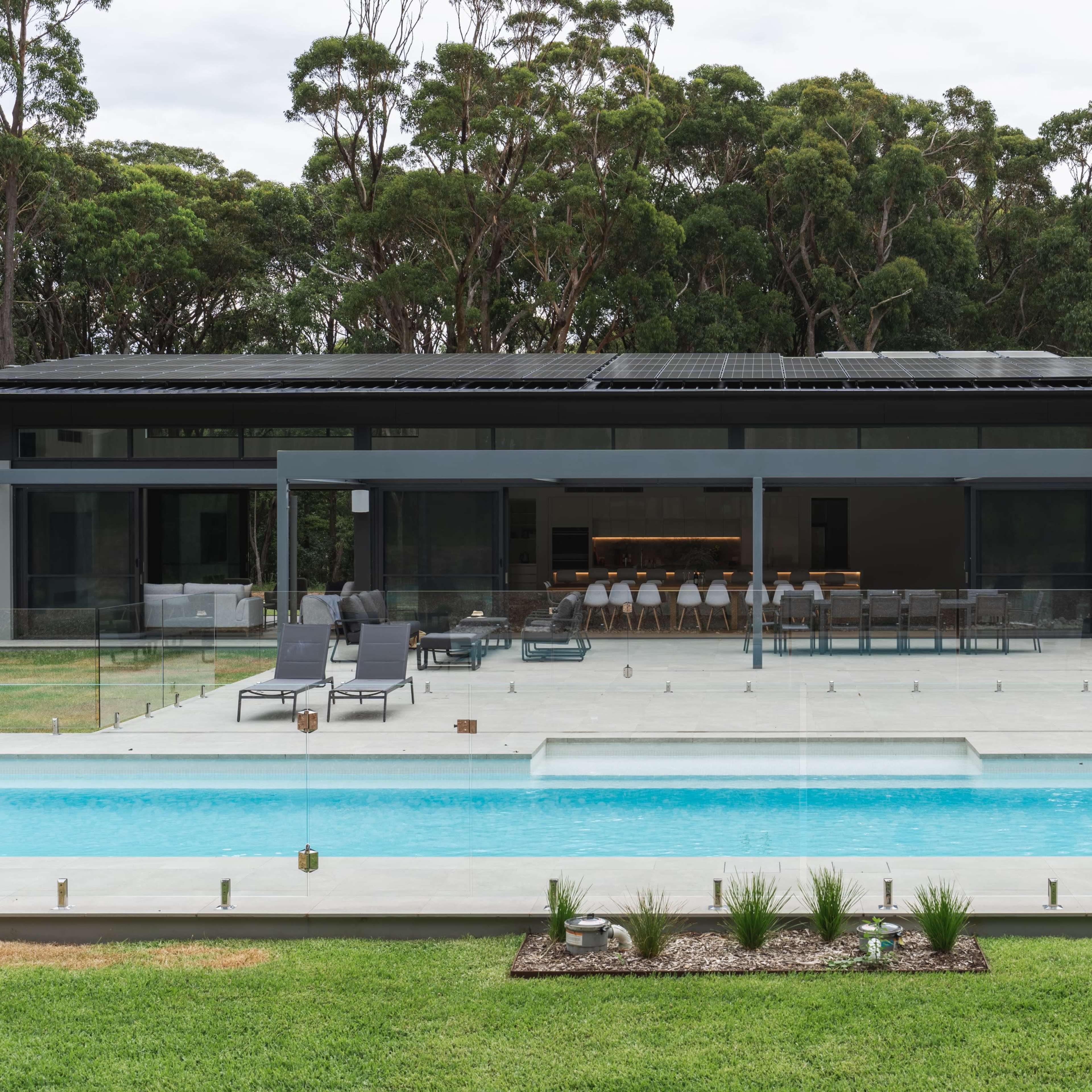 A modern home with a large glass-panelled back porch, overlooking a swimming pool surrounded by a manicured lawn and dense trees in the background.