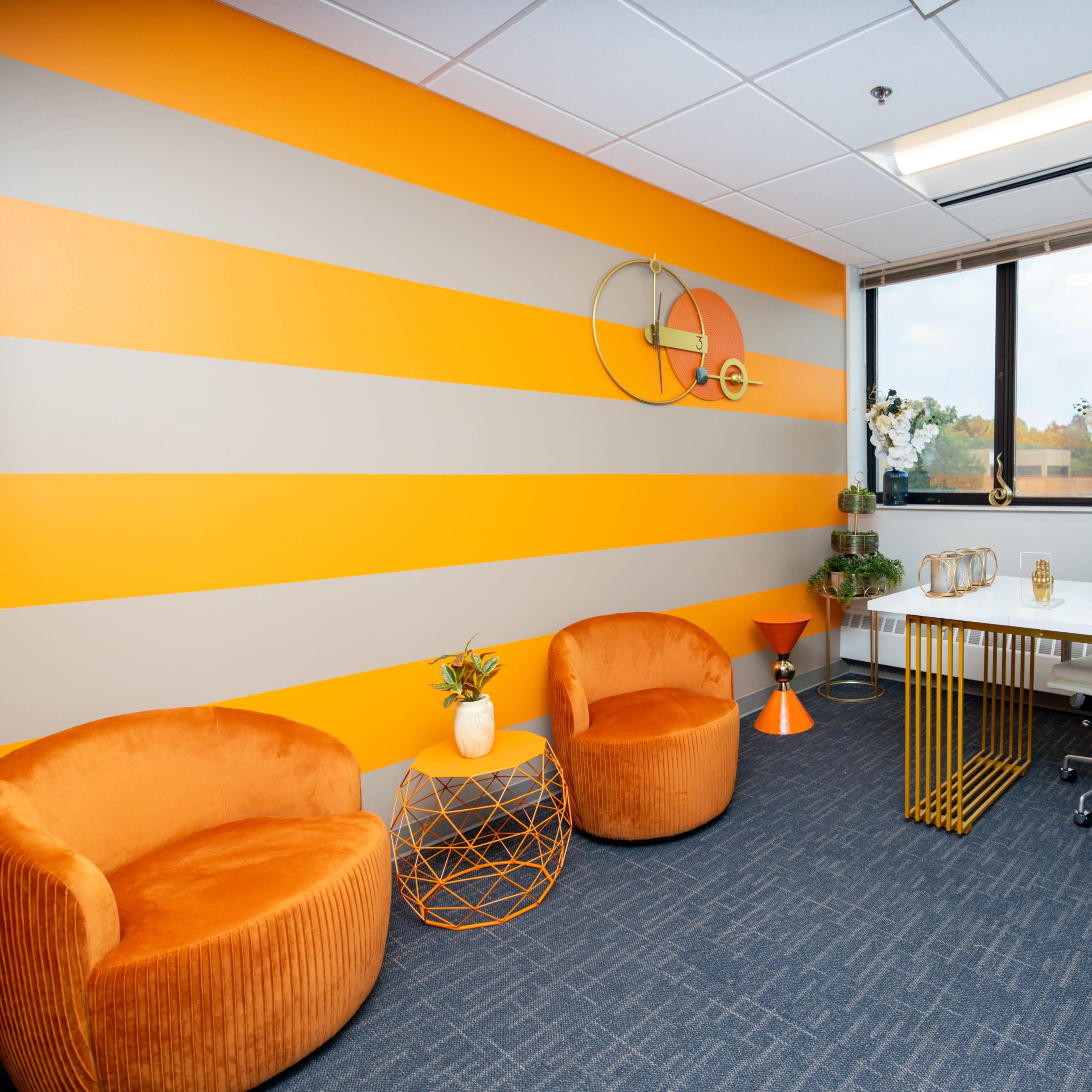 A modern office space featuring orange striped walls, two orange accent chairs, a white desk with a monitor, and decorative plants.