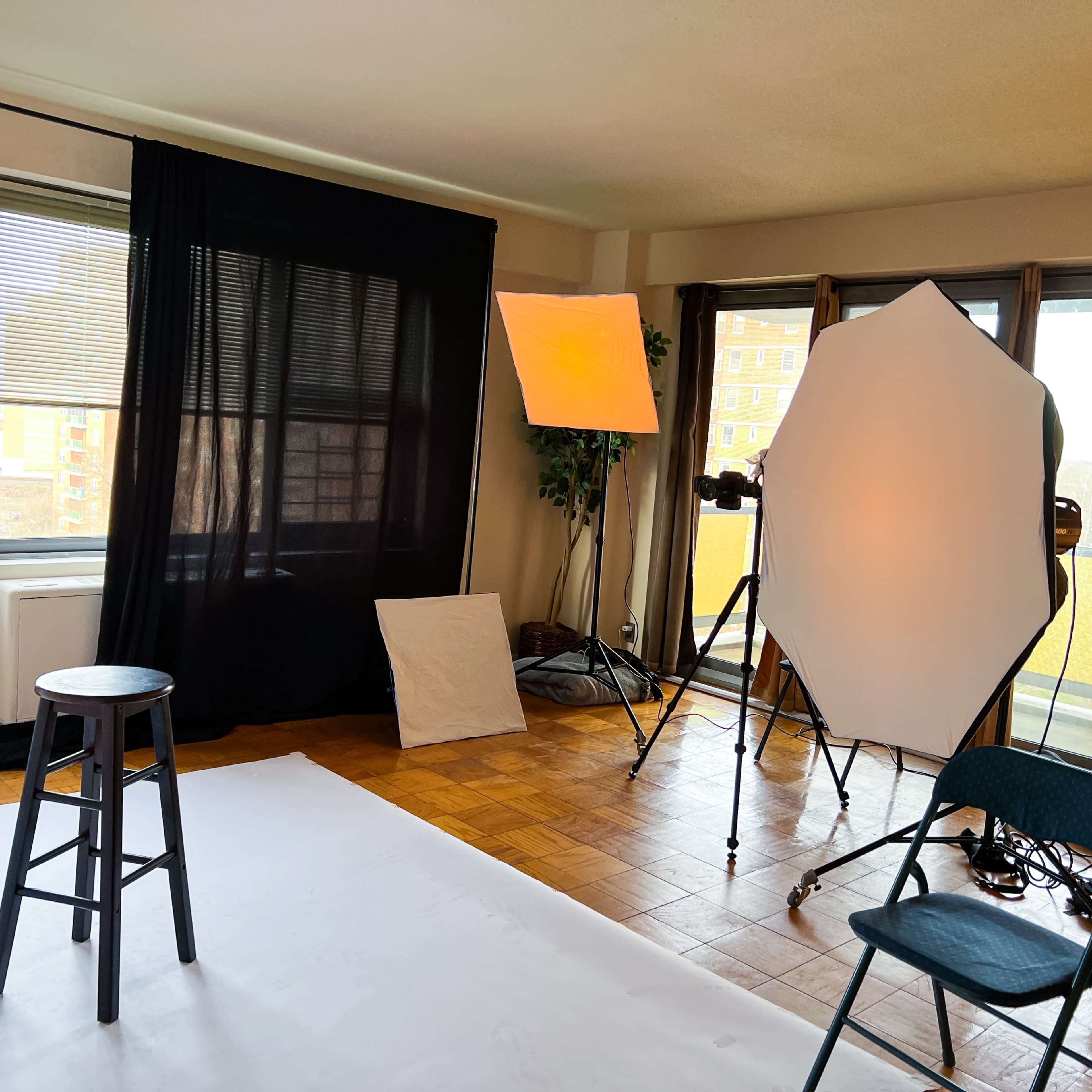 A well-lit photography studio setup featuring a black backdrop, a stool, and softbox lights arranged next to a large window.