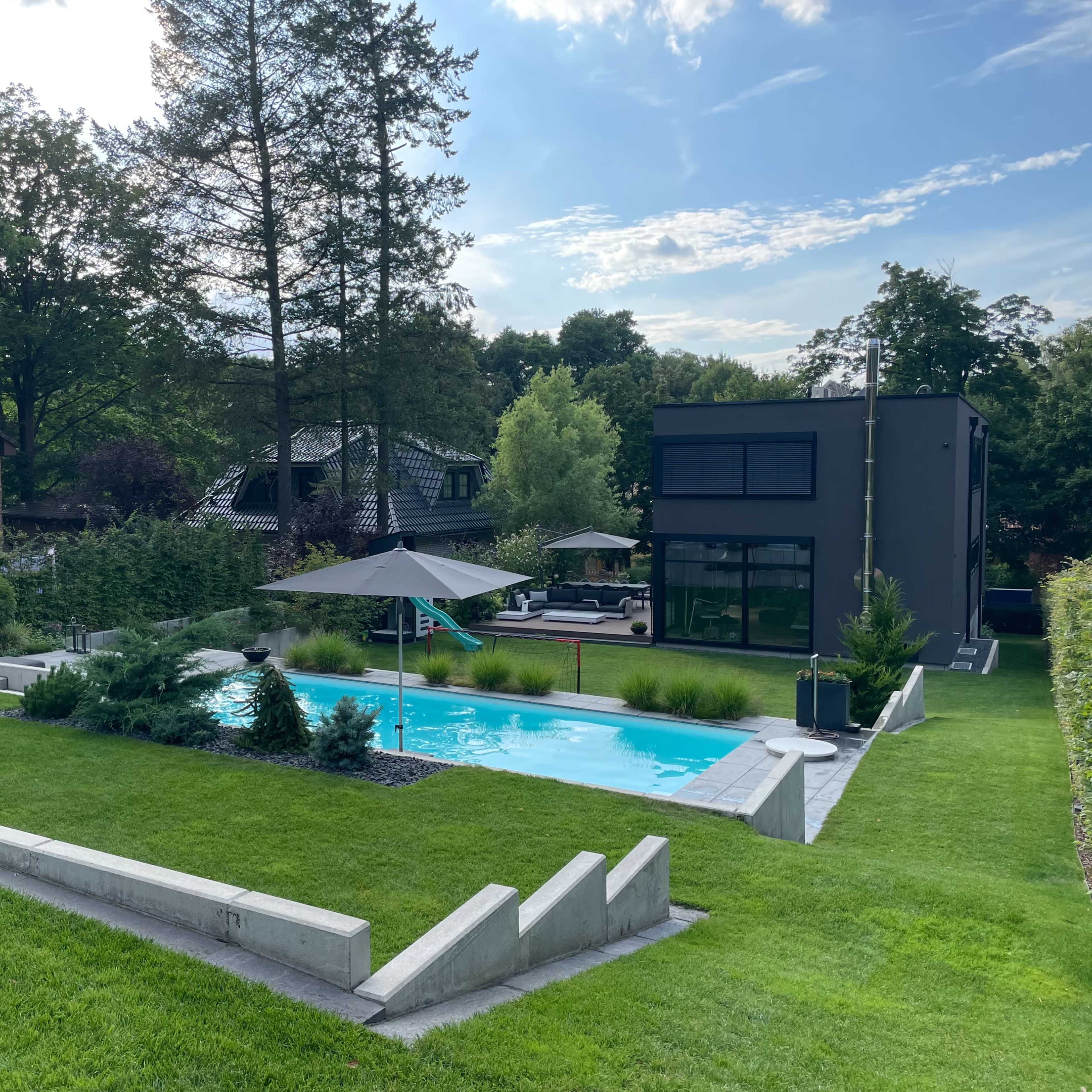 A landscaped backyard with a swimming pool, surrounded by green grass, trees, and a modern two-story building in the background.