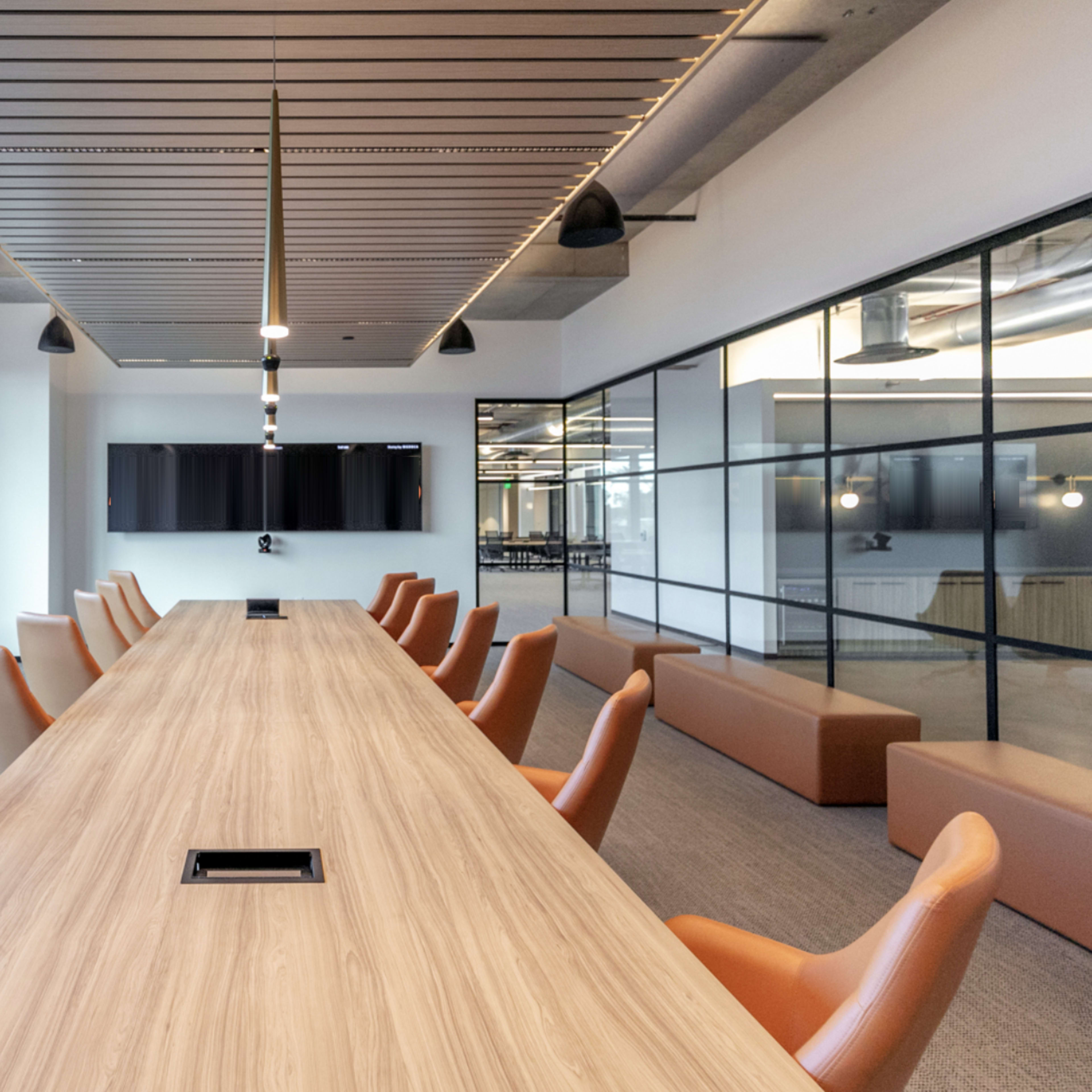 A long wooden conference table with orange chairs is set in a brightly lit meeting room featuring large windows and modern audio-visual equipment on the walls.