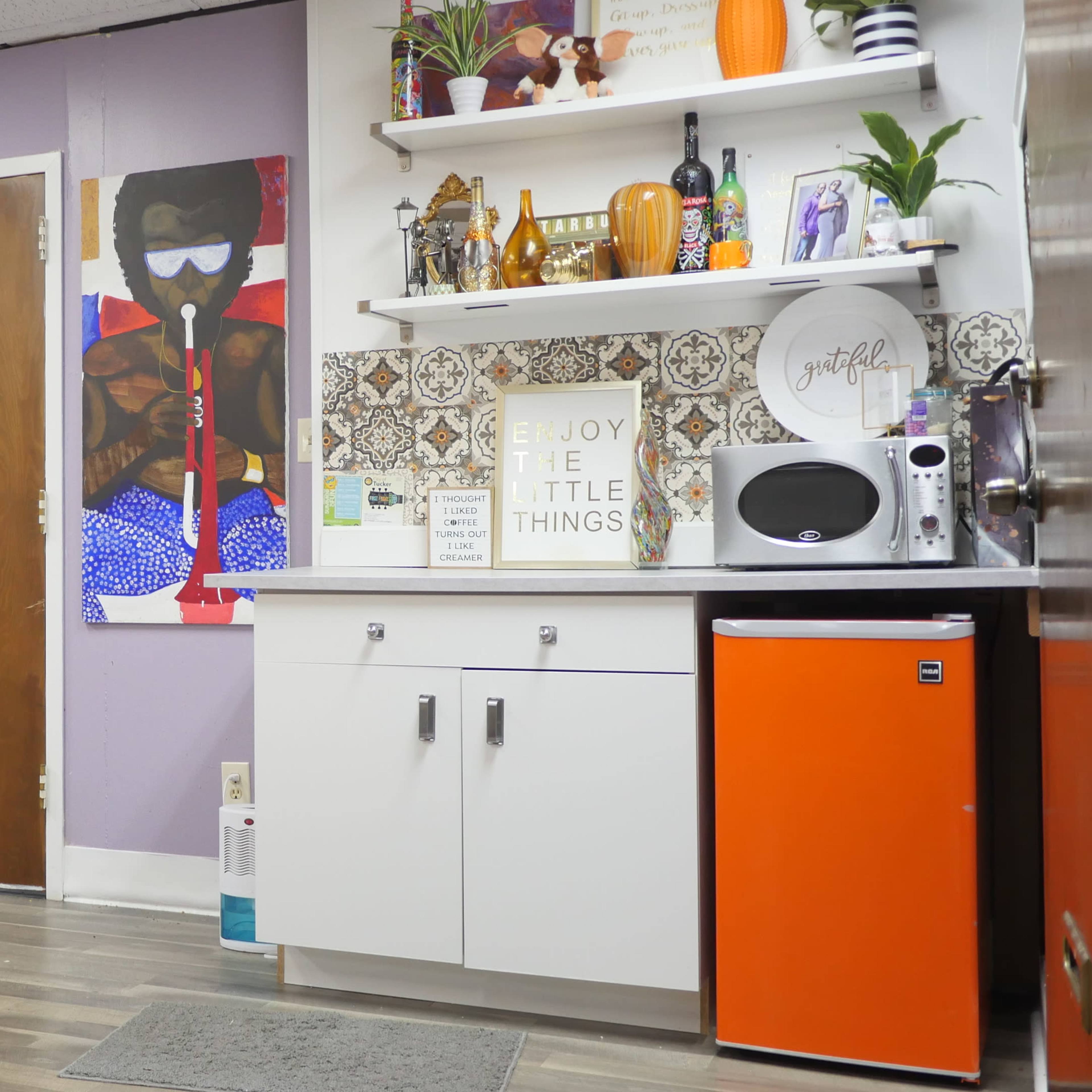 A room with purple walls, a small table, a kitchen area featuring a microwave, an orange mini fridge, and colorful wall art.