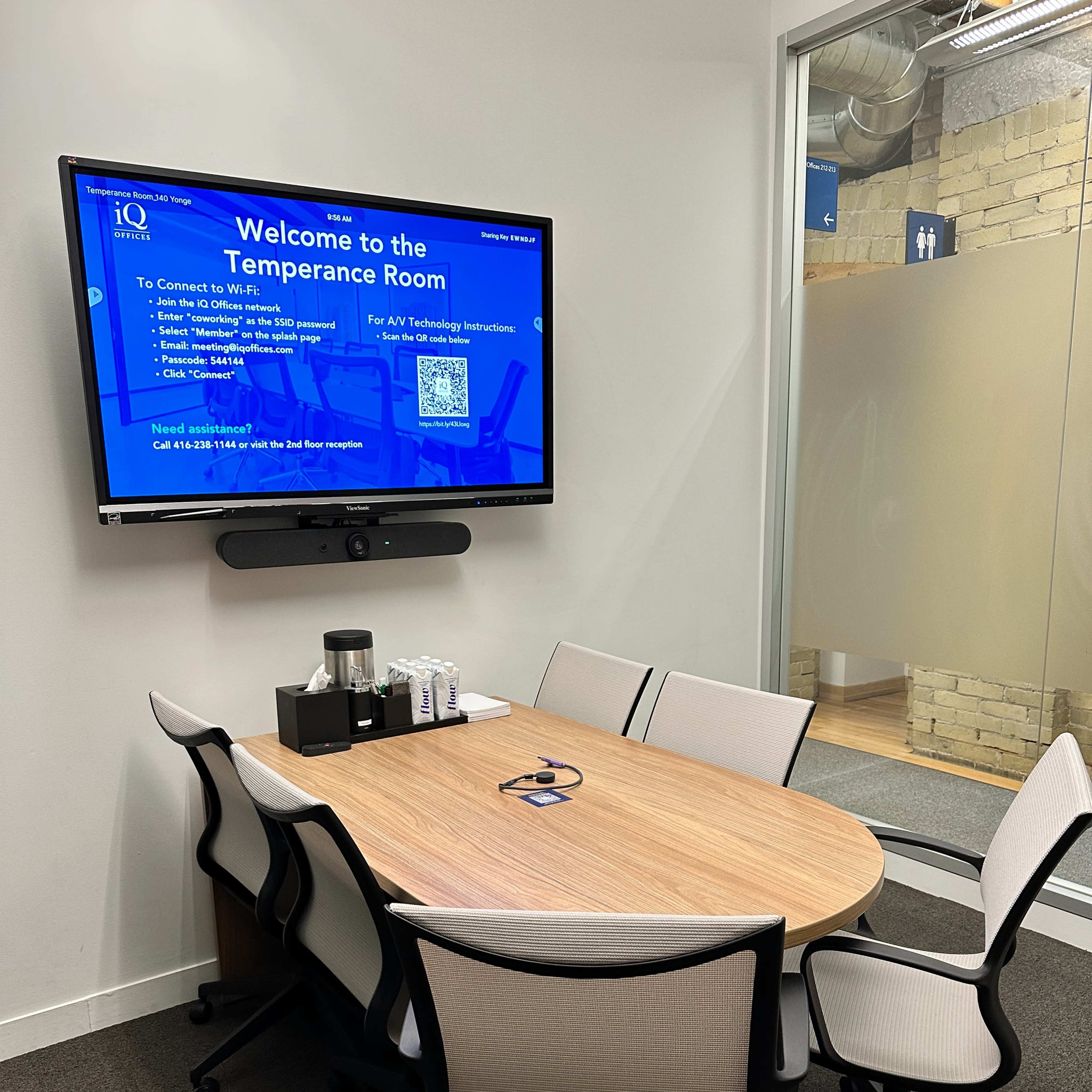 A small meeting room with a round wooden table and six chairs, a screen displaying "Welcome to the Temperance Room," and a coffee station on the table.
