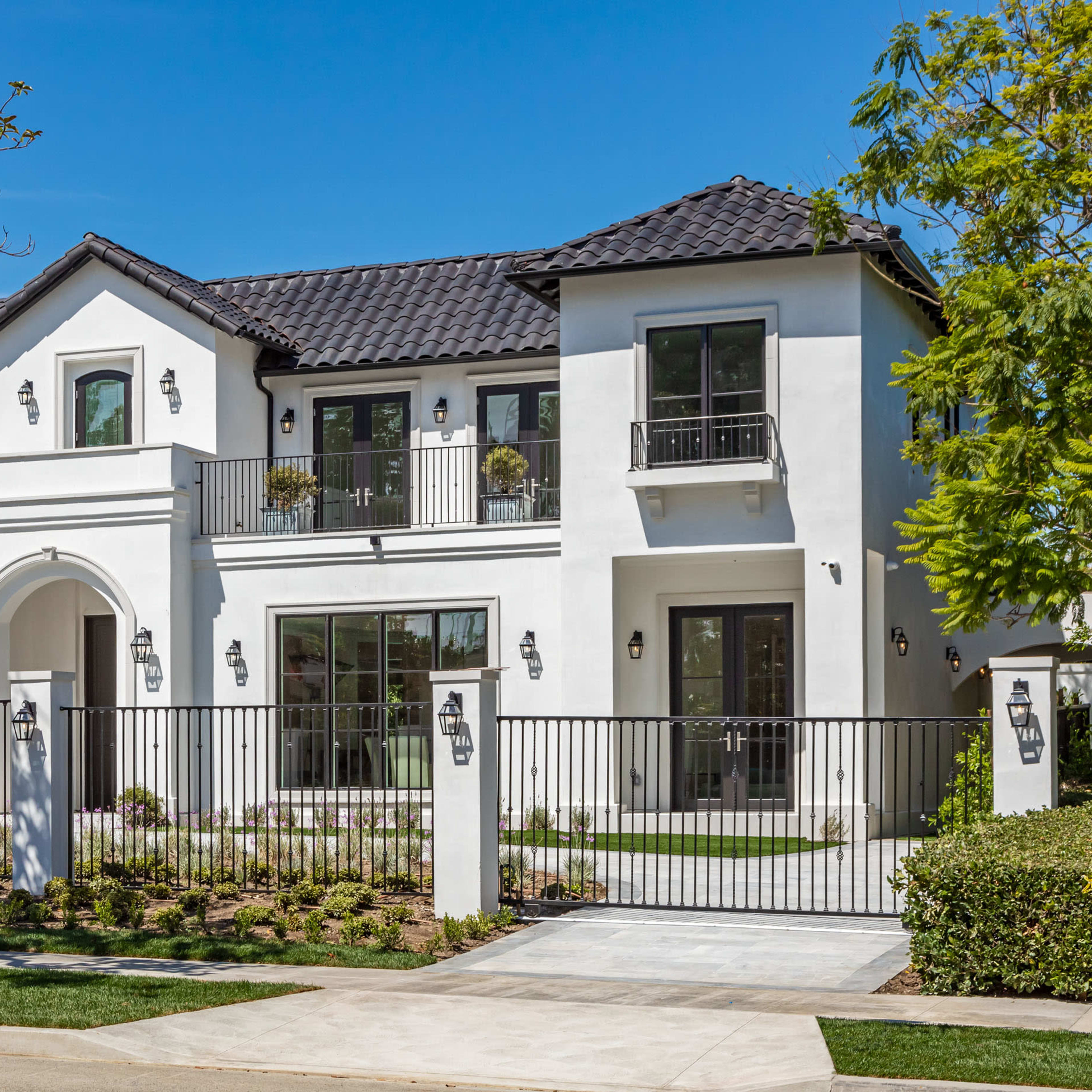 A large, modern white house with a tiled roof and black accents is enclosed by a black iron fence and features a landscaped front yard.
