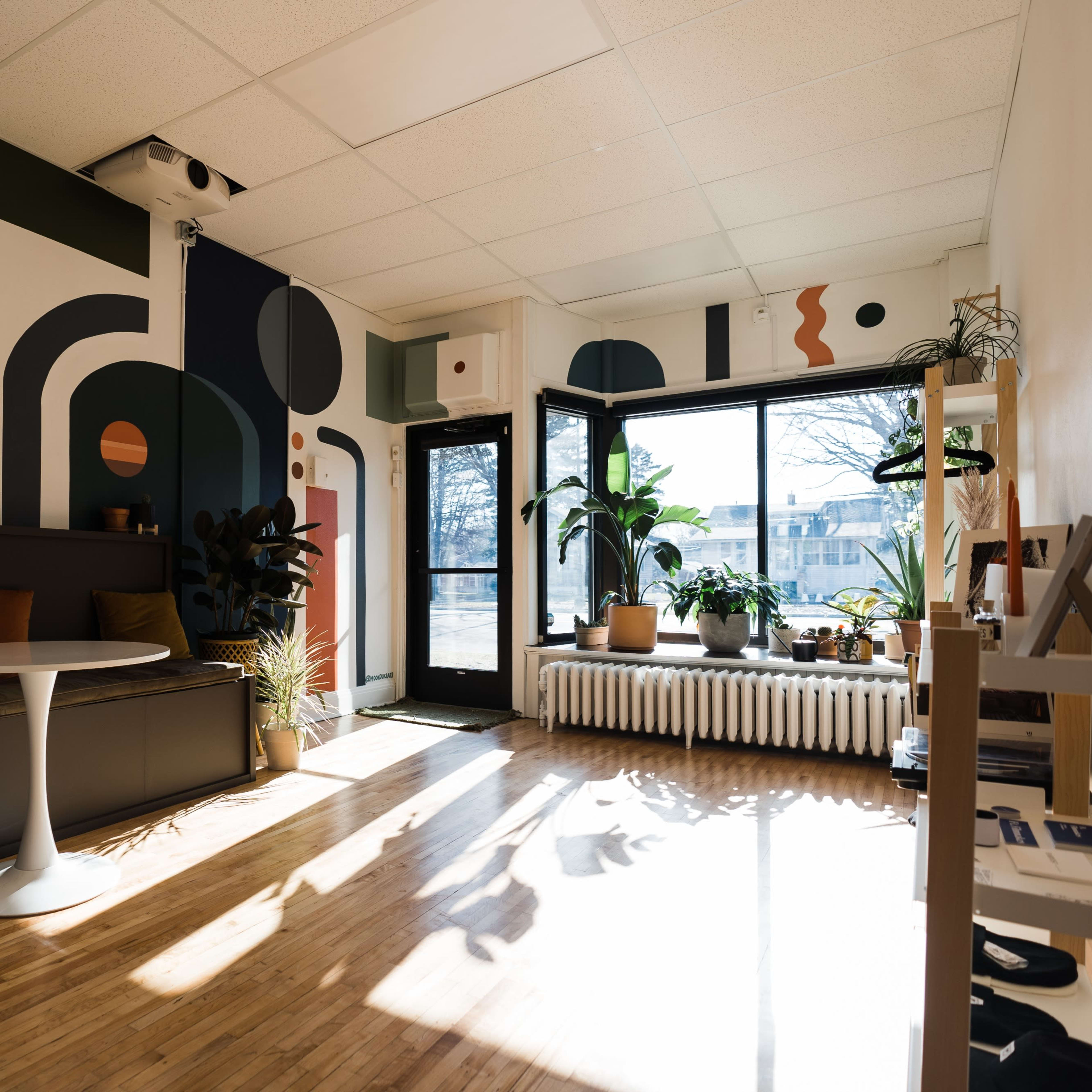 A modern interior space with a patterned mural on the wall, a round table, seating area, and large windows allowing natural light to enter.