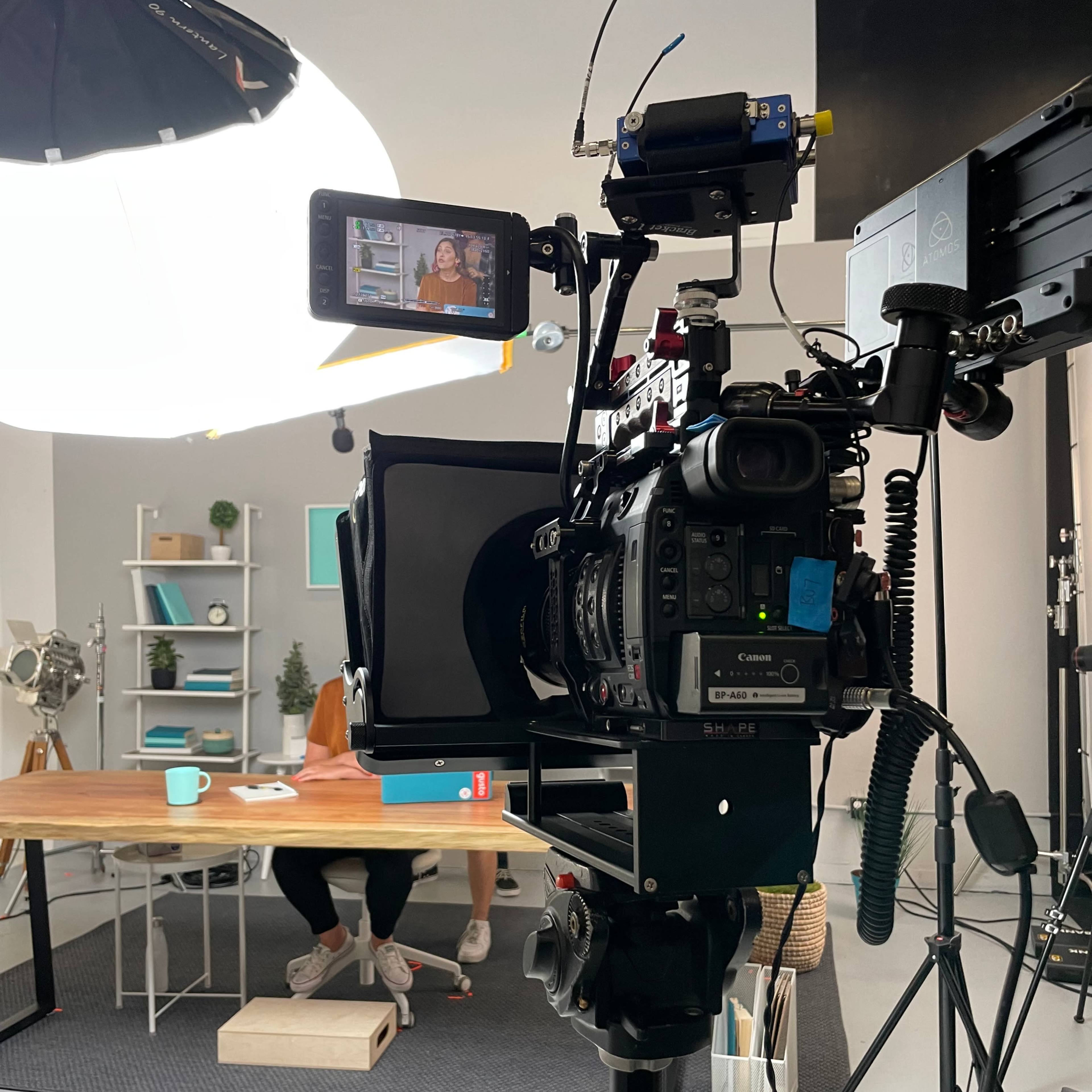 A video camera is positioned on a tripod, directed towards a woman sitting at a desk in a well-lit studio setup.