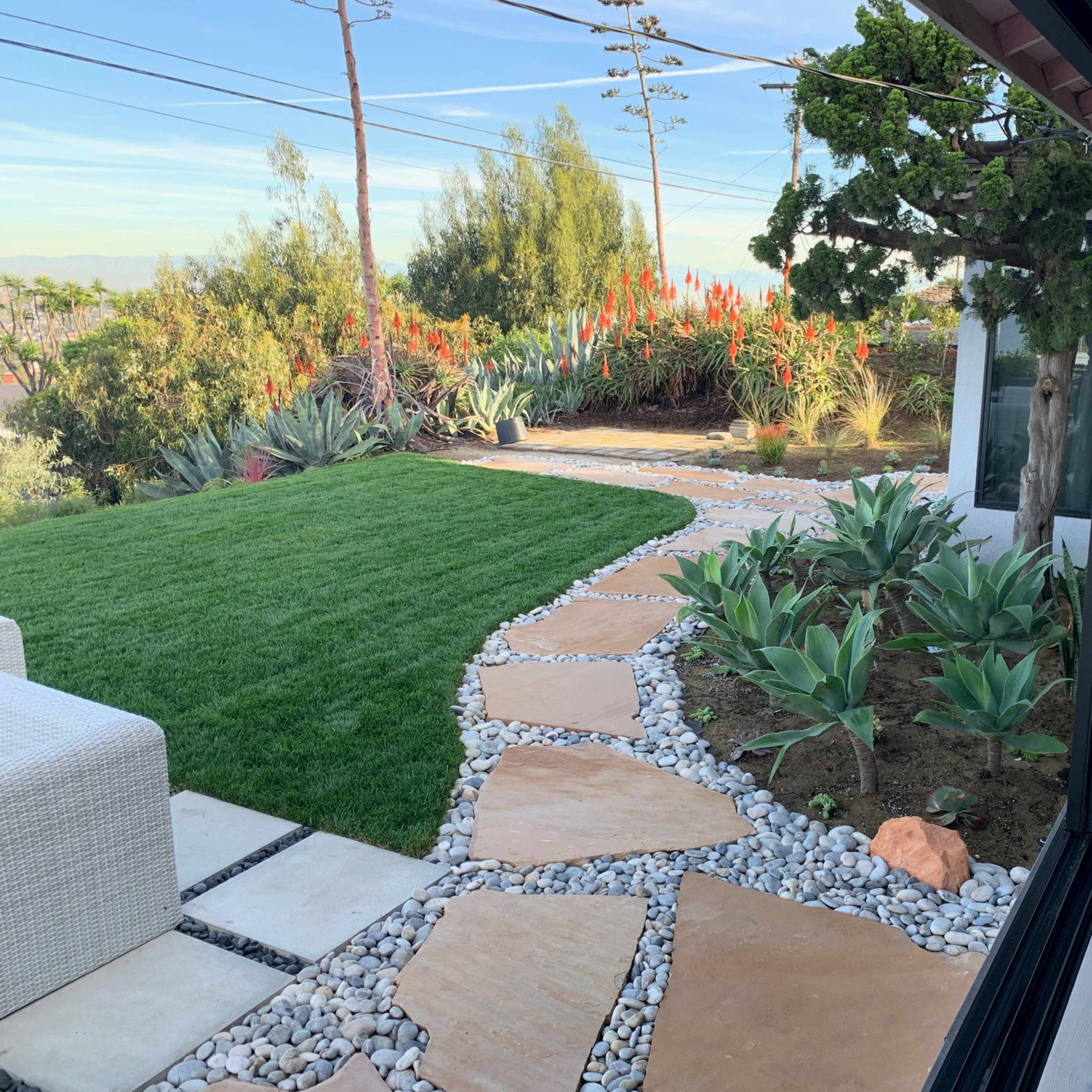 A landscaped garden with a stone pathway leading through green plants and a well-maintained lawn, framed by power lines and a distant tree line.