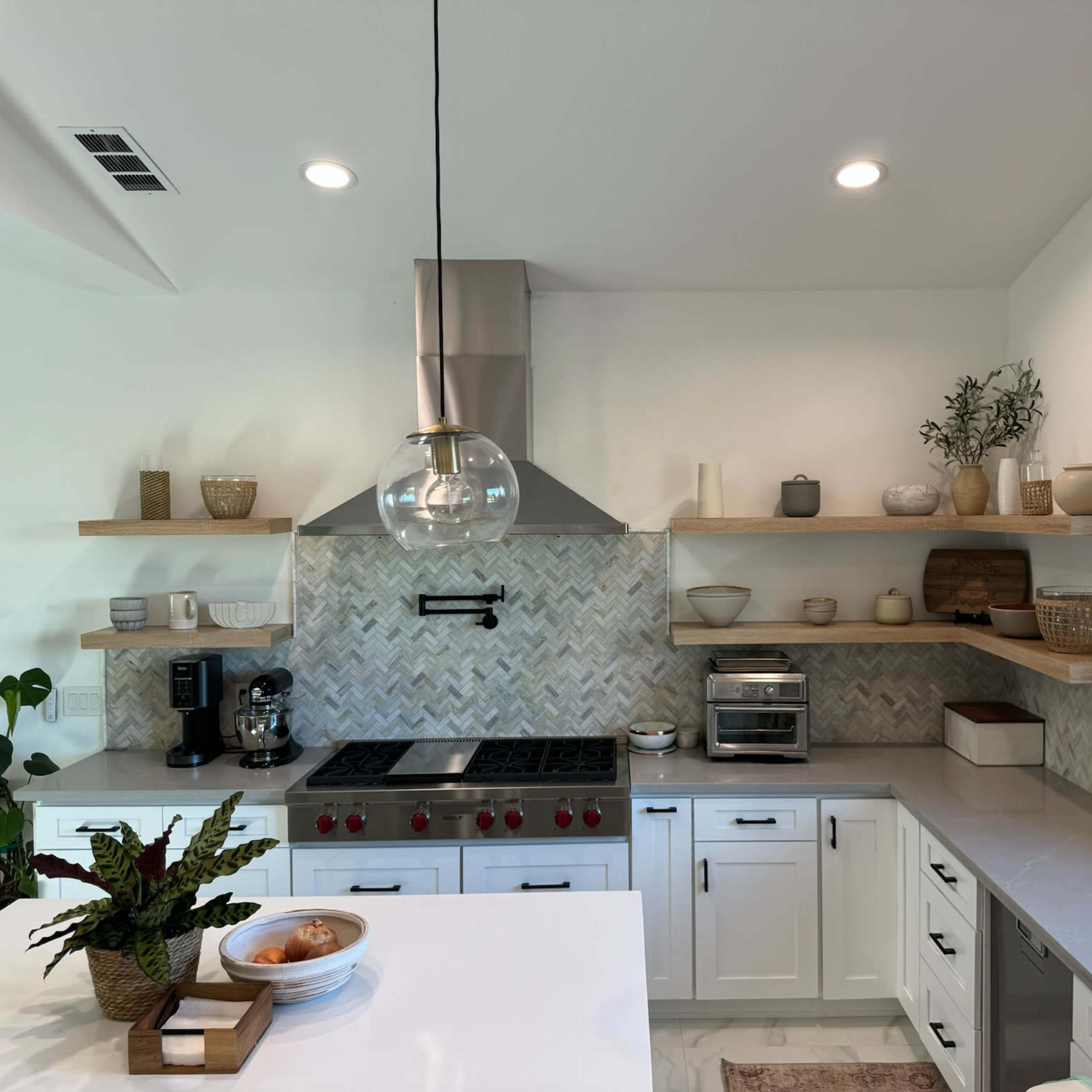 A modern kitchen features a stainless steel range hood, open shelving with decorative items, and a central island with a bowl of fruit.