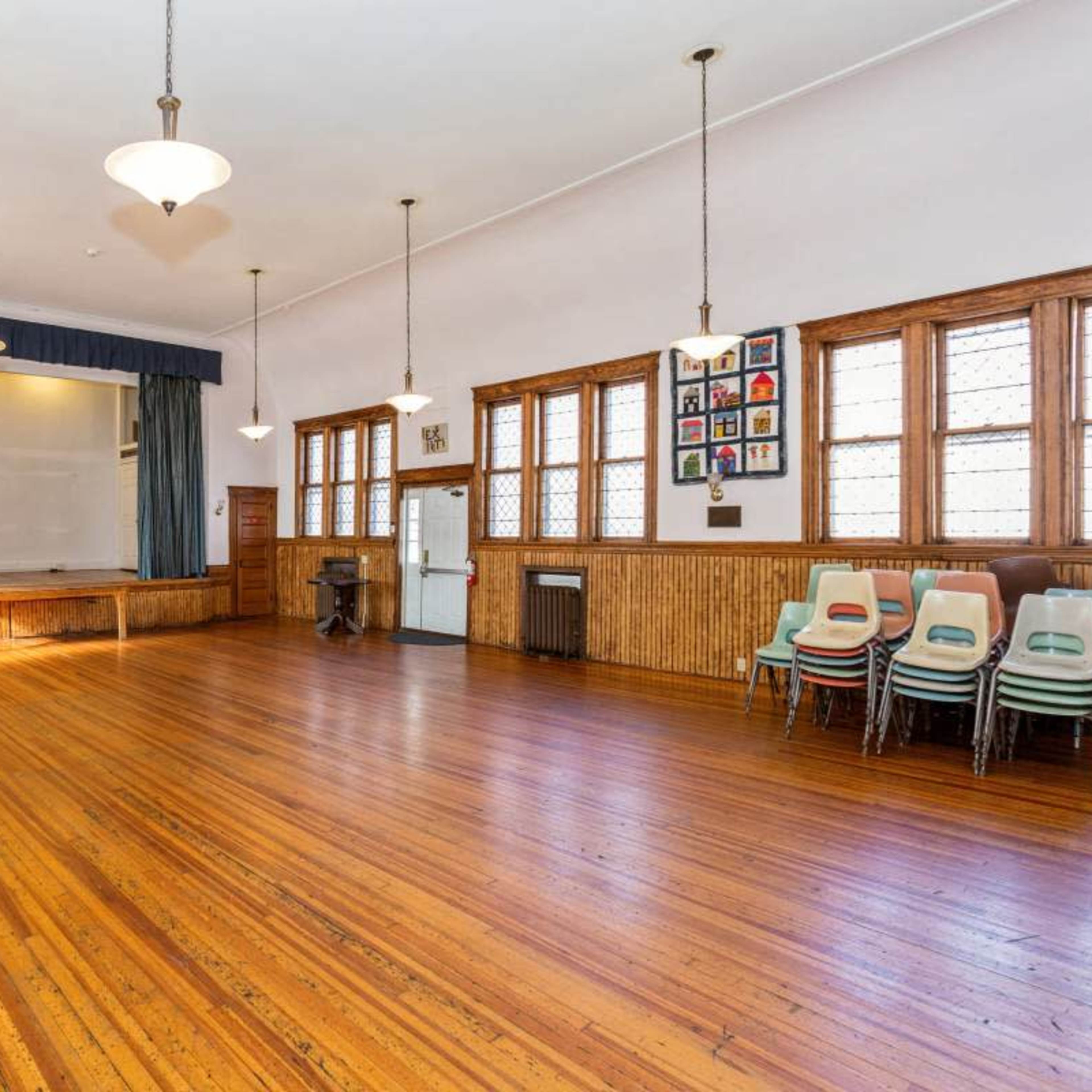 A large, empty room with wooden floors, tall windows, and chairs stacked against the wall.