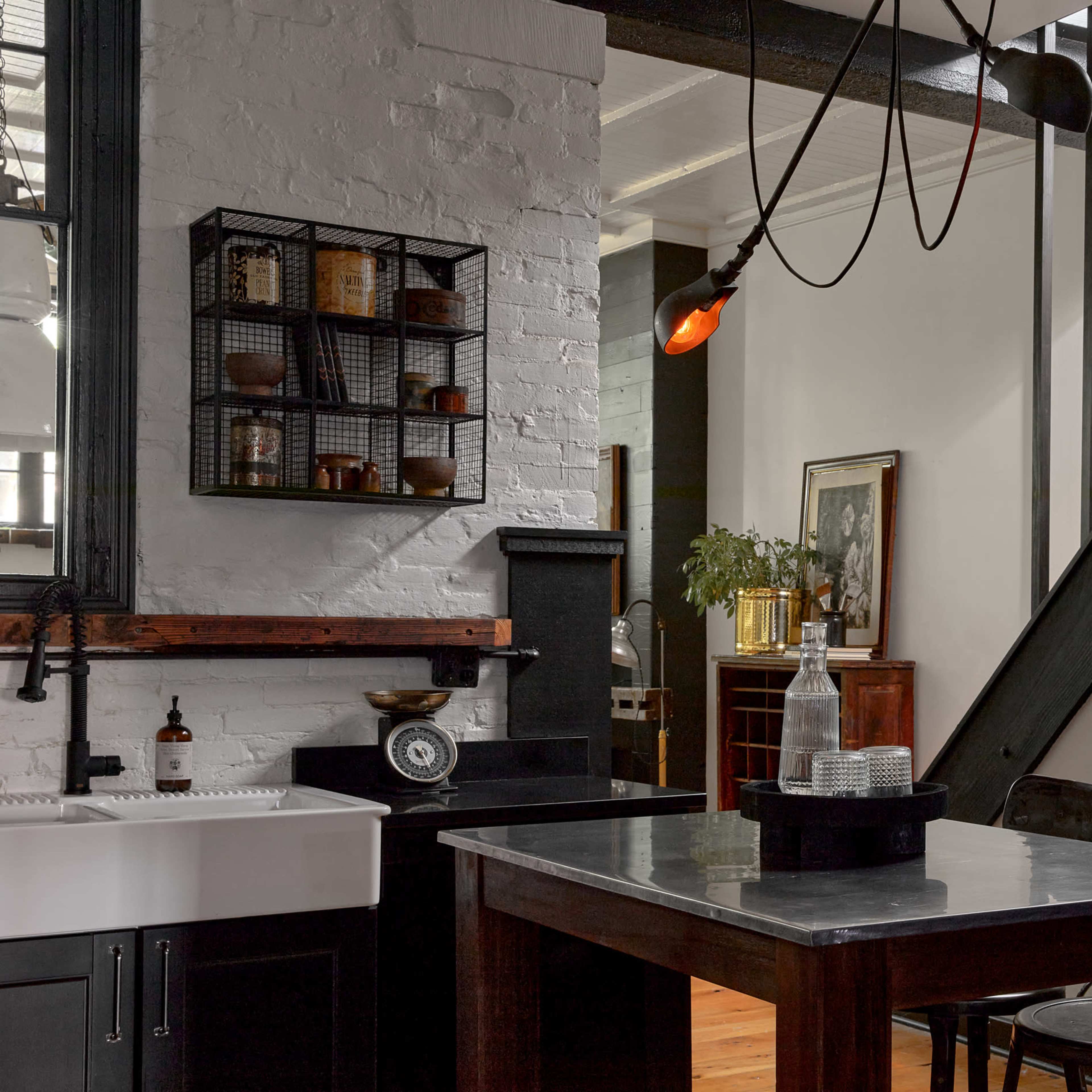 A modern kitchen with a large farmhouse sink, a marble island, and a metal shelving unit on a white brick wall, adjacent to a staircase and an entryway with a dark wooden door.