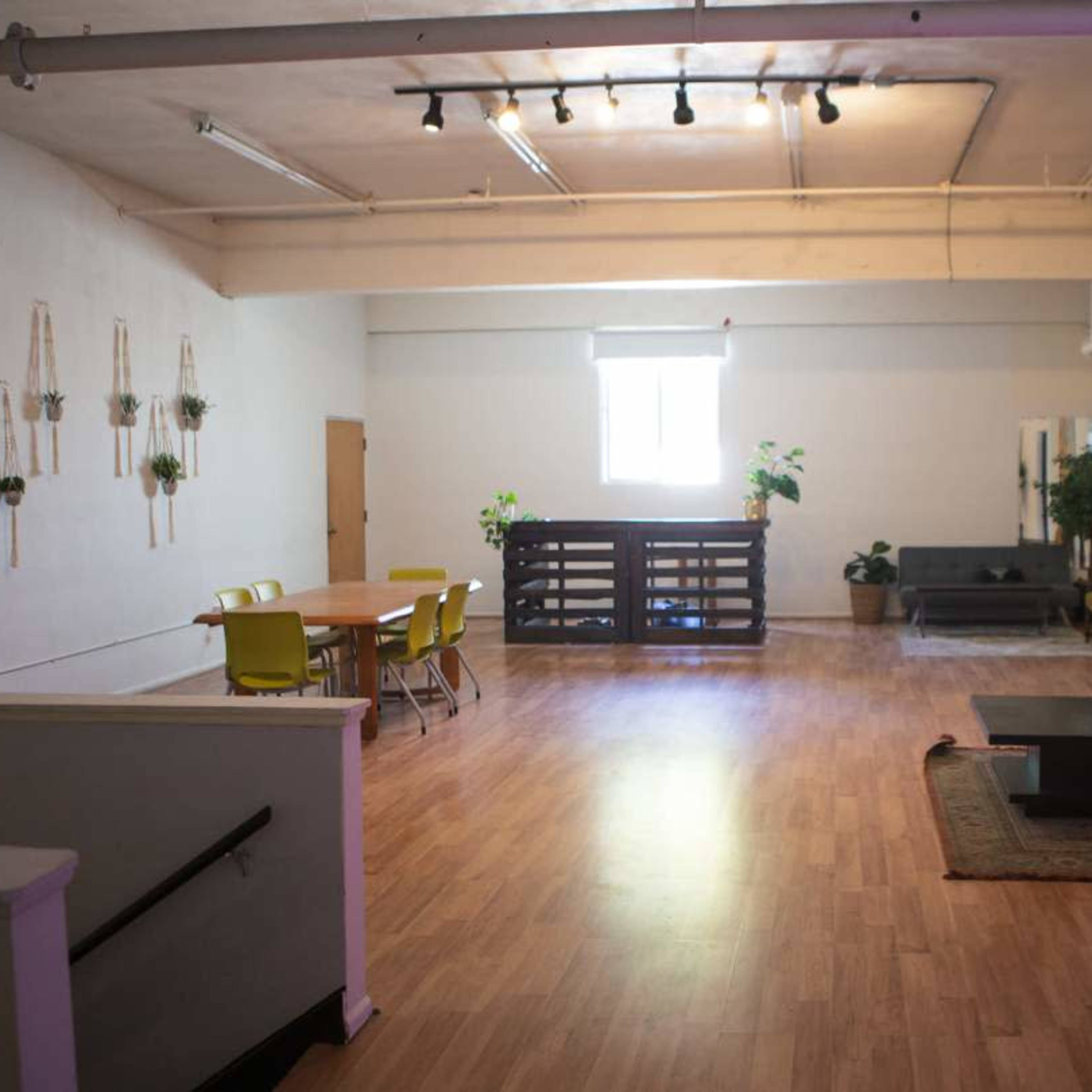 The spacious room features a dining area with yellow chairs, a lounge area with a gray sofa, and several plants arranged throughout the space.