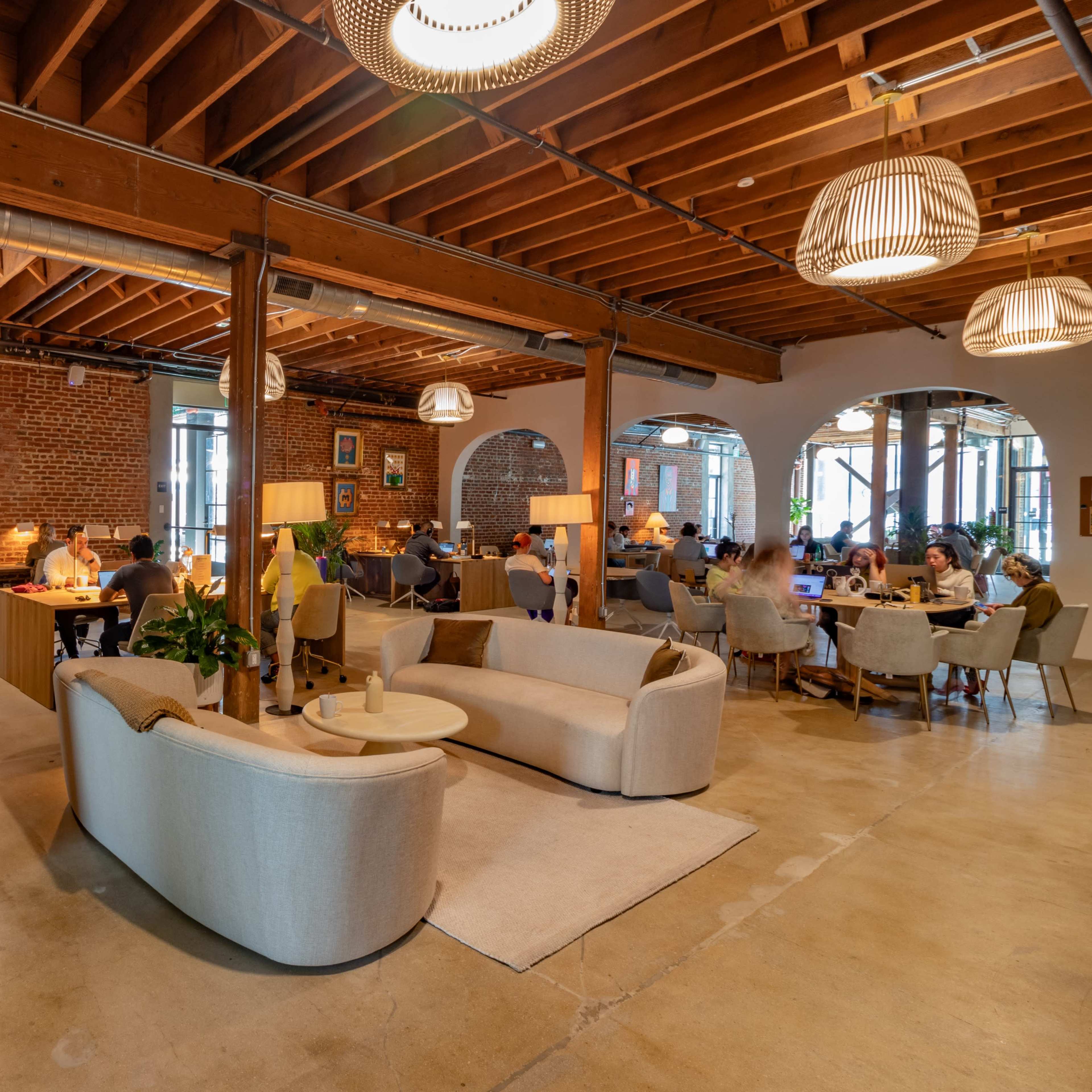 A spacious, modern café with wooden beams, brick walls, and comfortable seating areas filled with patrons engaged in conversation or working.