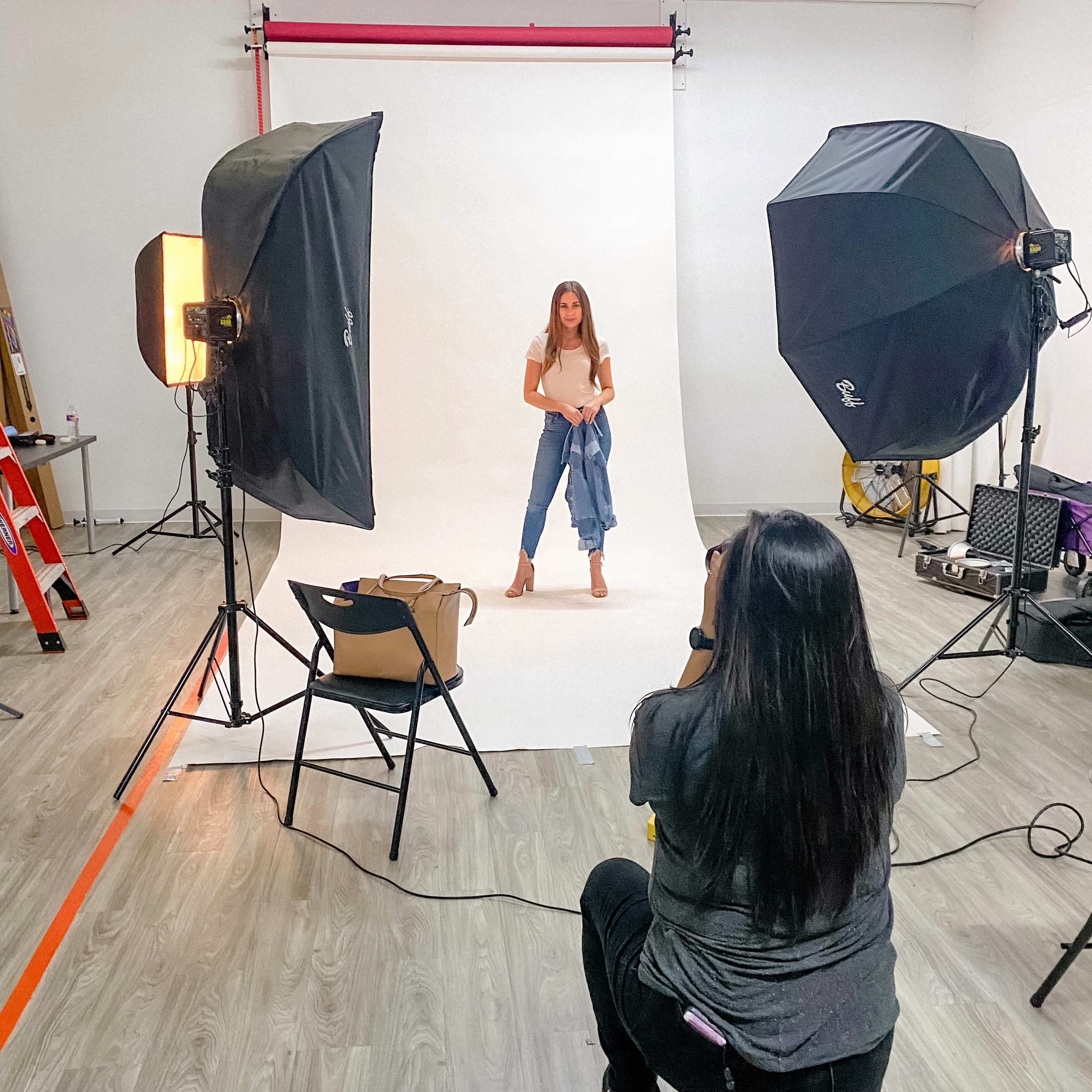 A photographer is capturing an image of a model posing in front of a backdrop in a studio equipped with lighting setups.