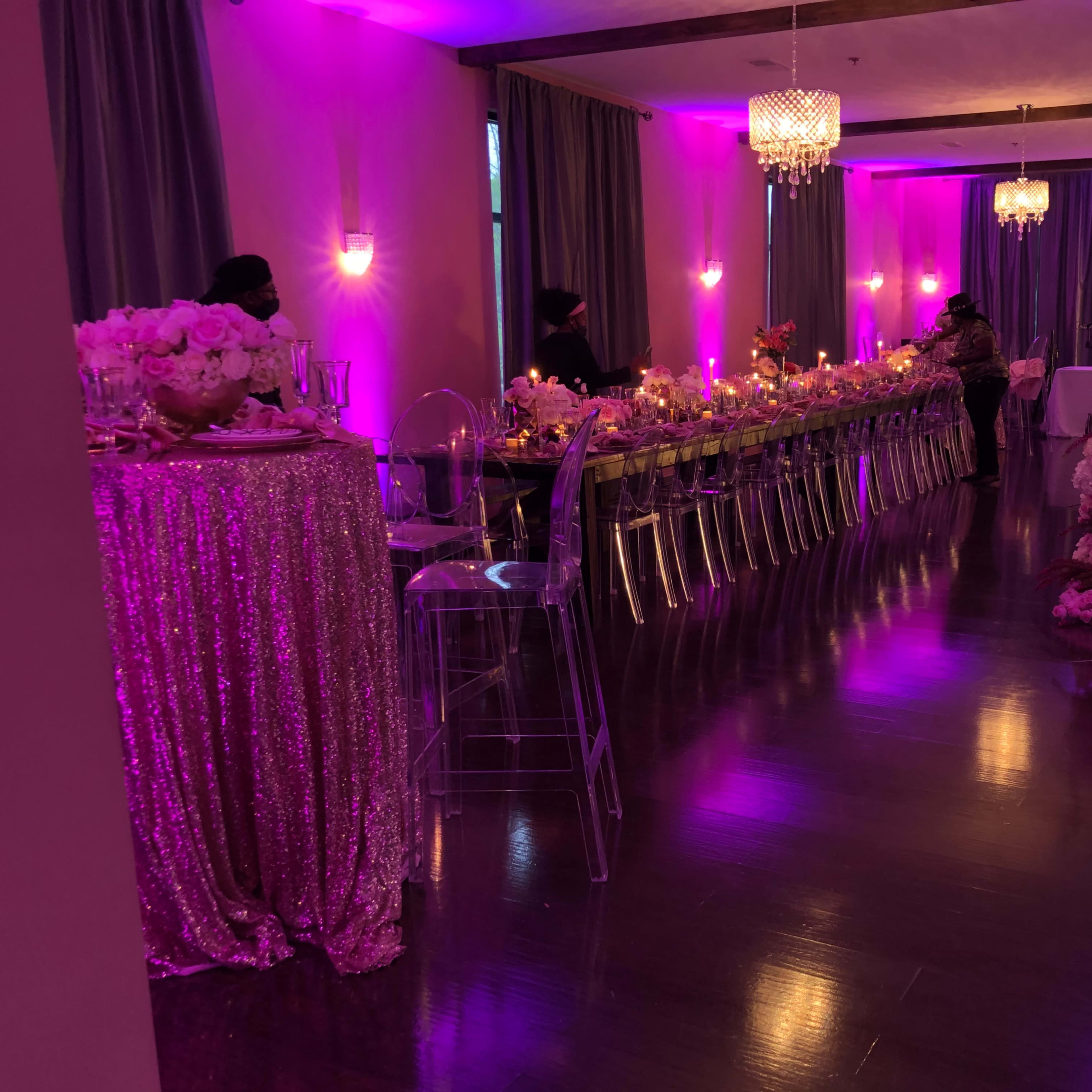 The scene shows a banquet hall with a long table set for a formal event, illuminated by purple lights and decorated with floral arrangements.