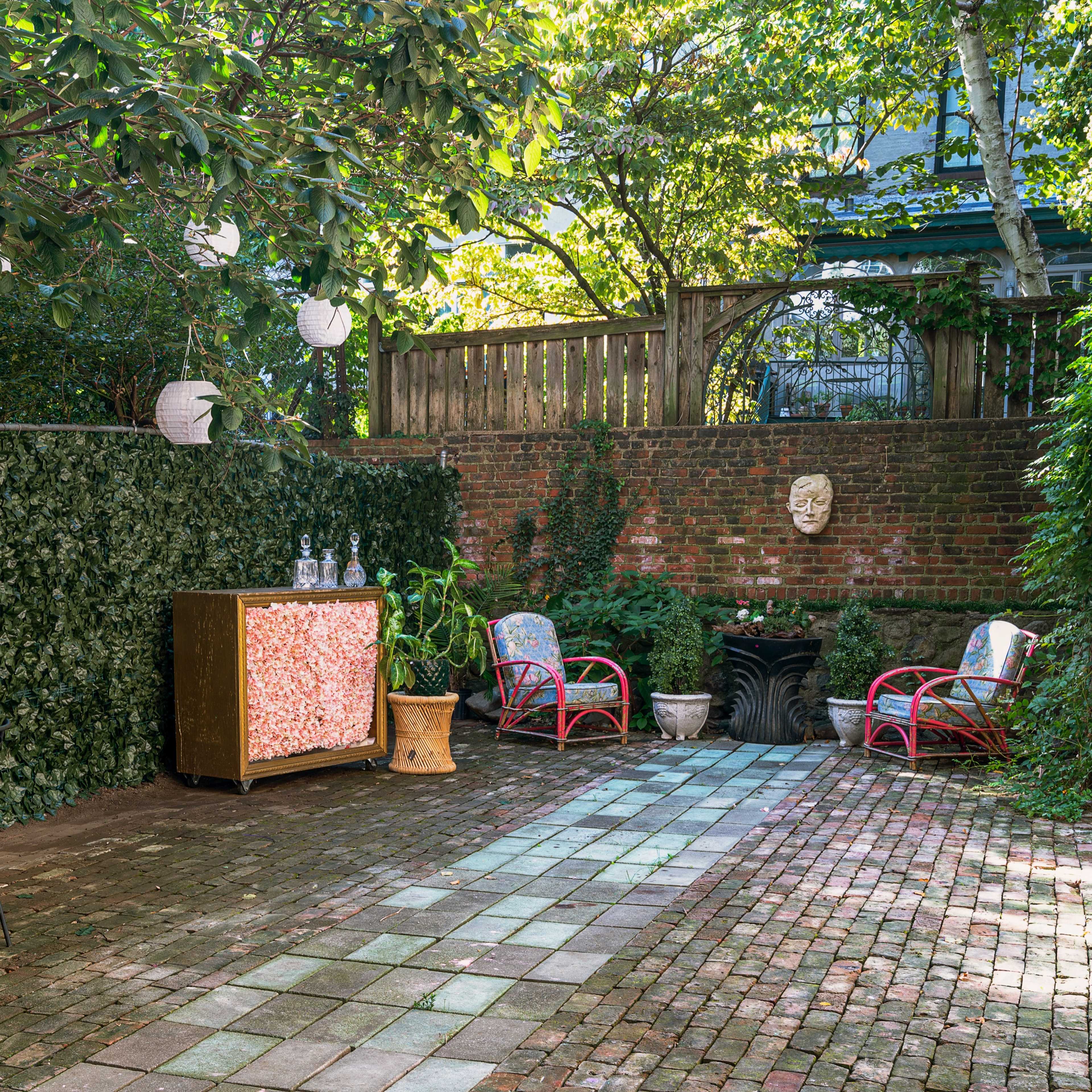 A shaded backyard garden features a stone patio with a decorative wooden cabinet, outdoor seating, and hanging lanterns amidst lush greenery.