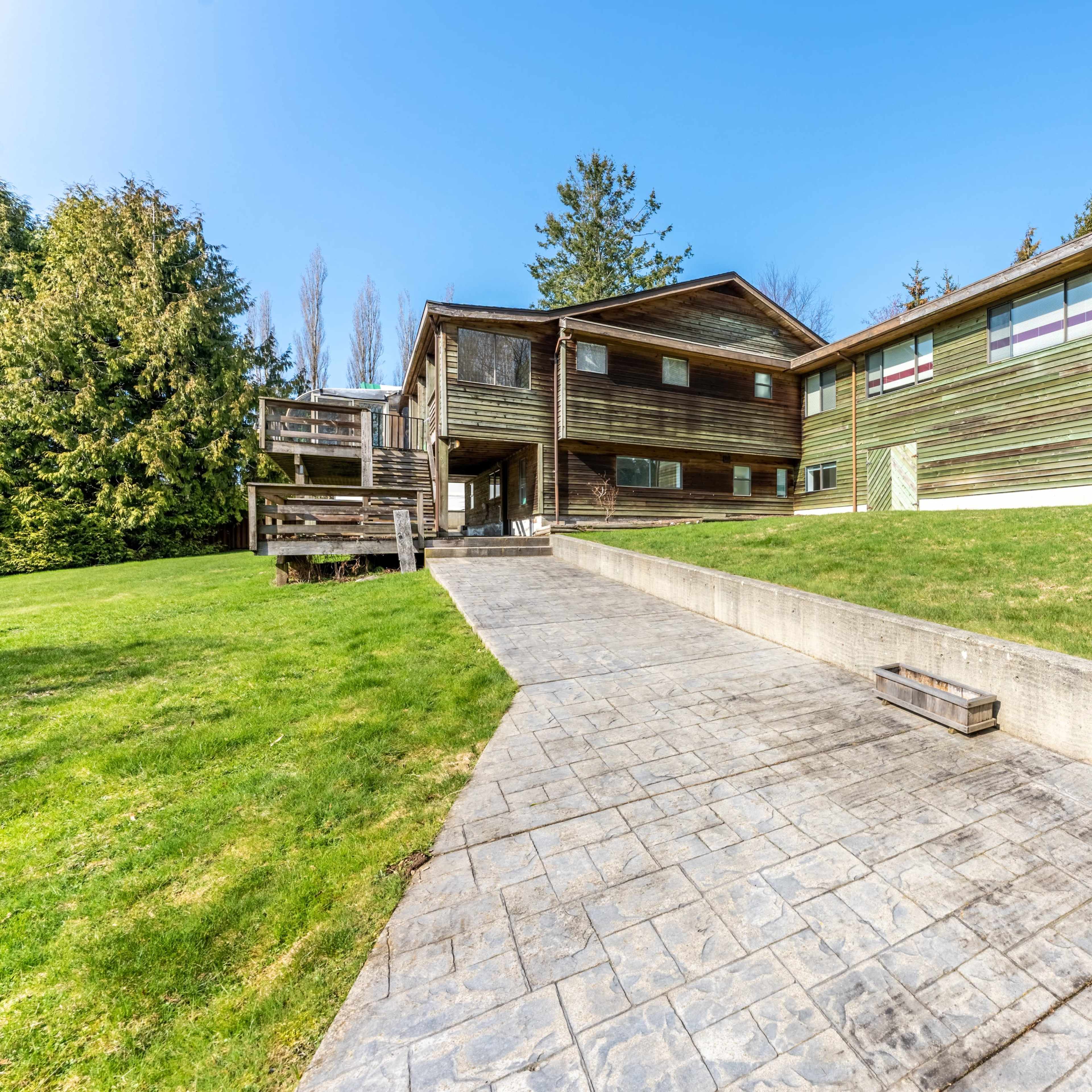 A large wooden house with multiple sections and windows is surrounded by a green lawn and paved pathway.