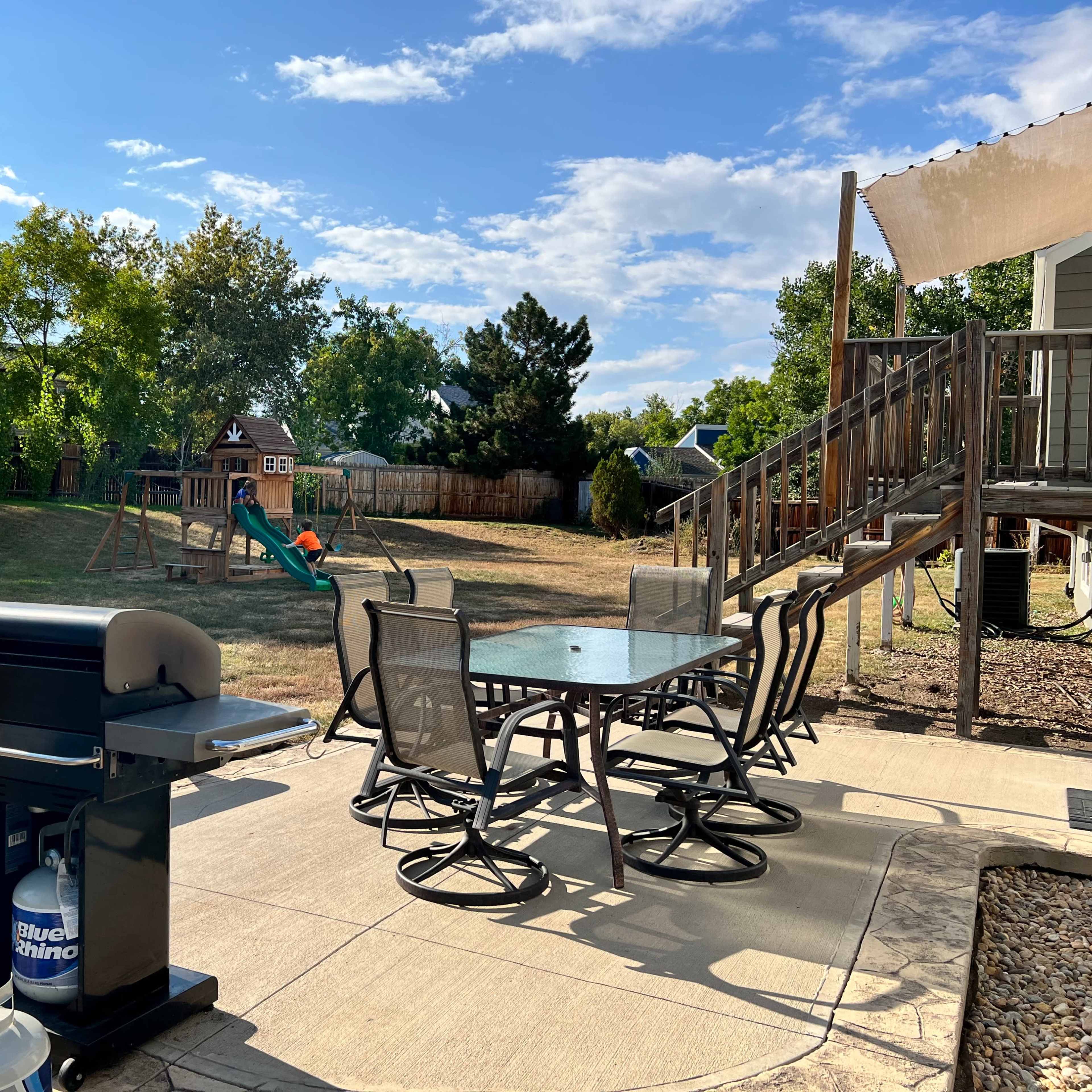 A backyard with a patio set, a grill, and a playground featuring a slide and swing set in the background.