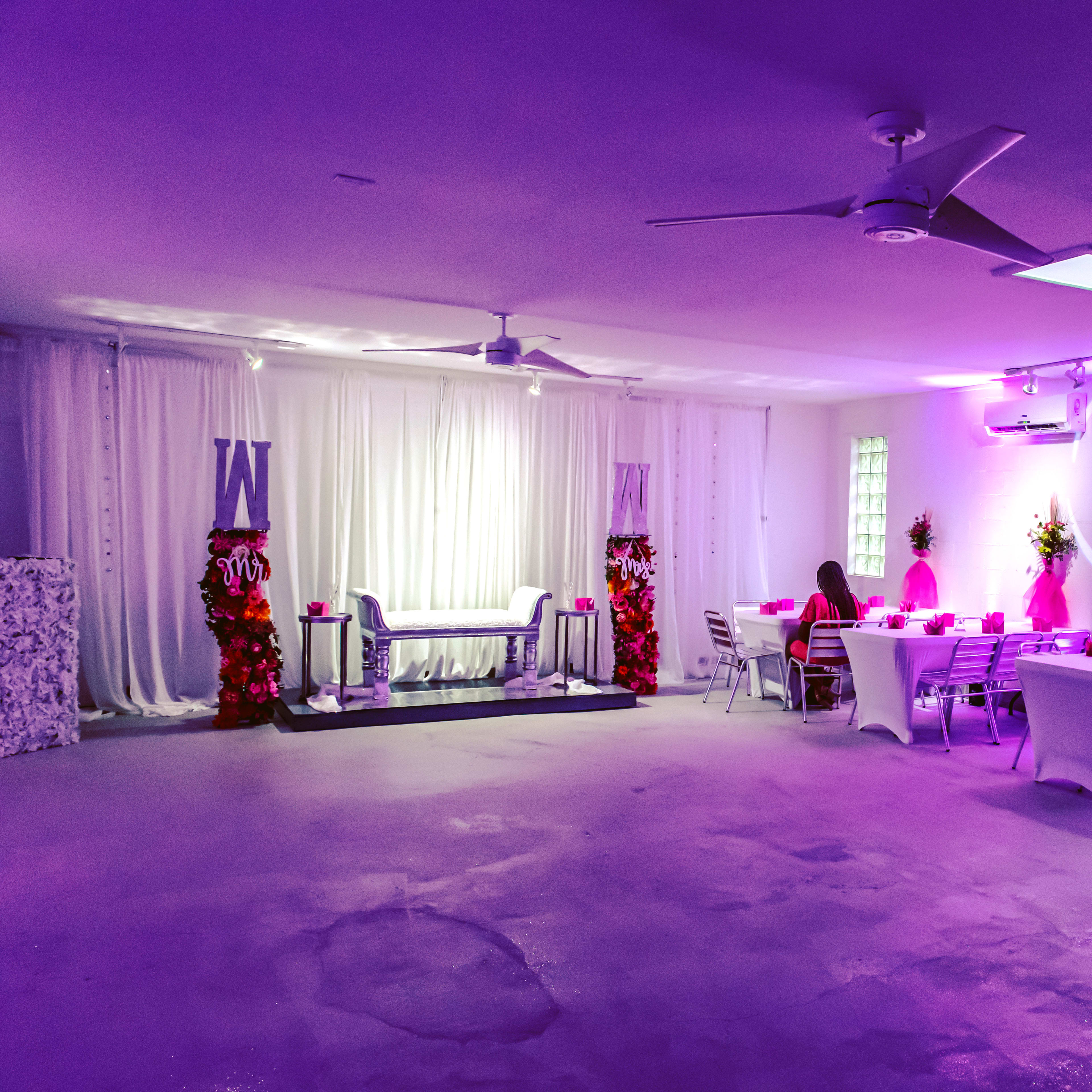 A partially illuminated event space with a decorated stage area, two floral arrangements, and a dining setup with chairs and tables.